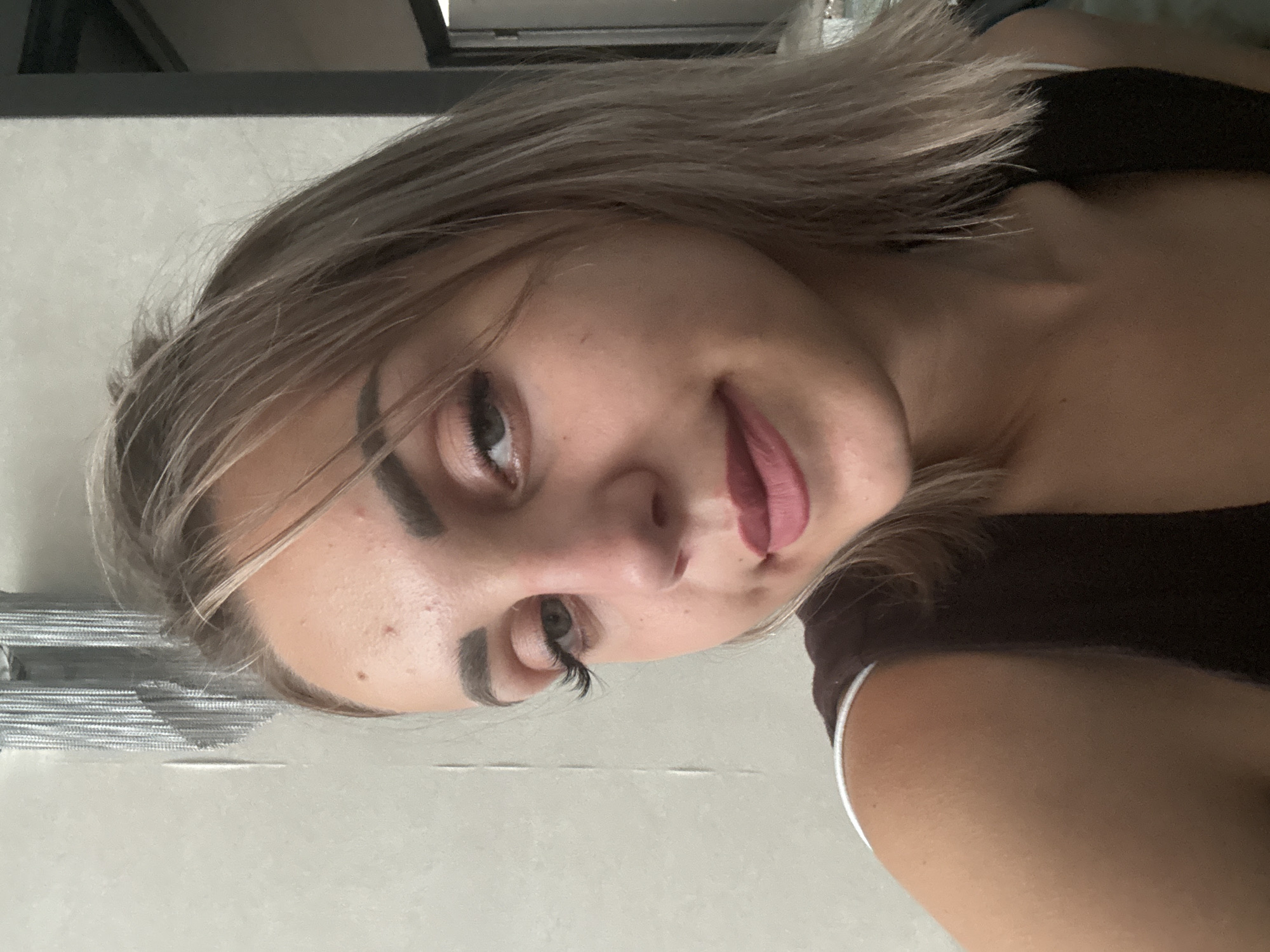 The image shows a young woman with blonde hair and light skin, wearing a dark brown top with white trim. She has on makeup, including dark eyeliner and mascara, and a deep pink lipstick. Her hair is shoulder-length and slightly tousled. She is looking towards the camera with her head slightly tilted. The background appears to be an indoor setting with a textured wall and a hint of a doorway in the upper right corner. There is no visible indication of her location in Yuzhno-Sakhalinsk, Russia.