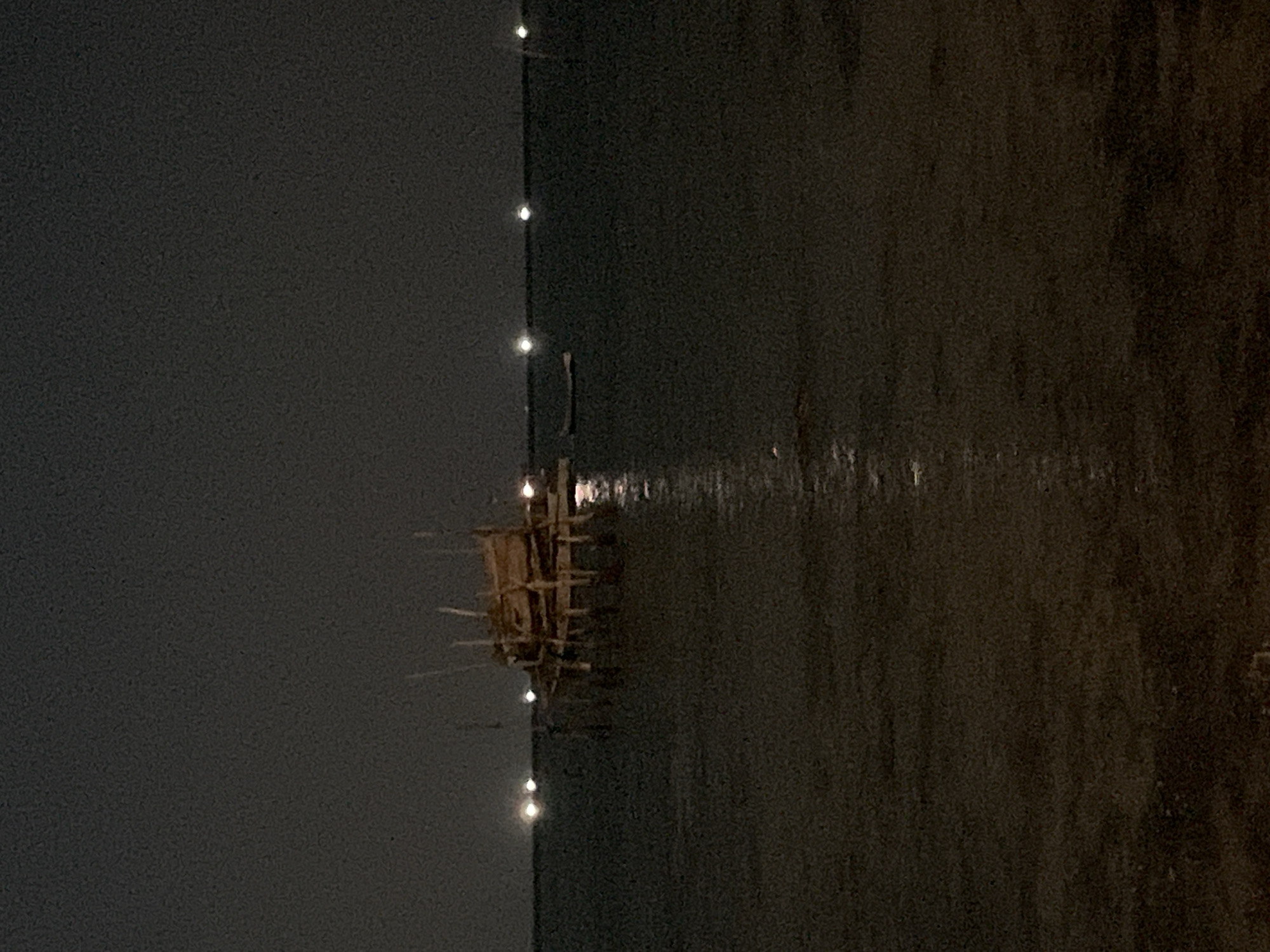 This image depicts a nighttime scene at Legonipah, Indonesia. The main subject is a wooden structure, possibly a fishing platform or a simple hut, standing in dark water. The structure is made of branches and sticks, with some protruding outwards. It appears to be illuminated by several small, bright lights that are also reflected in the water below, creating a shimmering effect. These lights seem to be strung along a pole or line extending from the left side of the frame into the water, creating a vertical line of illumination.
The water itself is dark and calm, reflecting the minimal light sources. The background on the left is a very dark, smooth surface, possibly a wall or a shoreline, also faintly illuminated by the distant lights. On the right, the water transitions into a textured, darker mass, possibly representing a continuation of the water or a different area of the environment. The overall impression is one of a quiet, possibly remote, and dimly lit setting. The time of day is clearly night, given the darkness and the artificial lighting. There is no visible text in the image. The scene evokes a sense of tranquility and perhaps a touch of mystery due to the low light conditions and the solitary nature of the structure.