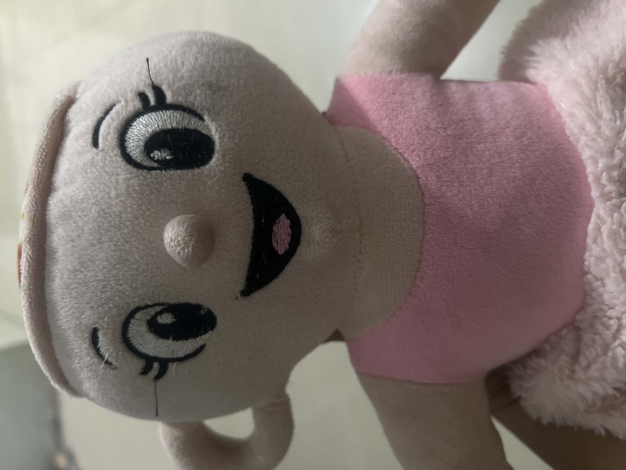 A close-up, waist-up shot shows a plush doll that resembles a tea cup with a face. The doll has a light tan, rounded head with embroidered features including large, black eyes with white highlights, thin black eyebrows, a small nose, and a wide smile with pink stitching inside the mouth. The doll has a single ear on its left side and is wearing a pink leotard and a fluffy pink tutu. The background is blurred and appears to be an indoor setting with a light-colored wall. The image does not provide any information about the city of Klapanunggal, Indonesia, or any people.