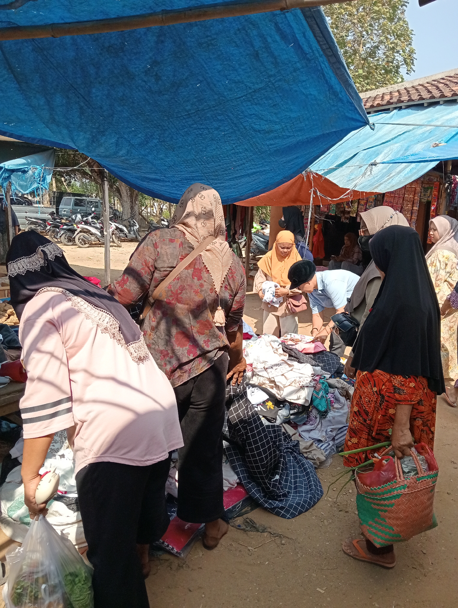 In Gunung Sugih, Indonesia, an outdoor market bustles with activity under a makeshift canopy of blue and orange tarps. Several people, mostly women dressed in hijabs and modest clothing, are browsing through piles of garments displayed on the ground and on tables. One woman in a pink shirt and black pants holds a plastic bag filled with green vegetables and a white object, possibly a computer mouse. Another woman, in an orange hijab, is shown holding a bundle of white fabric. A man in a light blue shirt is bending down, seemingly examining the merchandise. The ground is dirt, and in the background, motorcycles and a silver van are parked. The scene captures a typical day at a local Indonesian market.