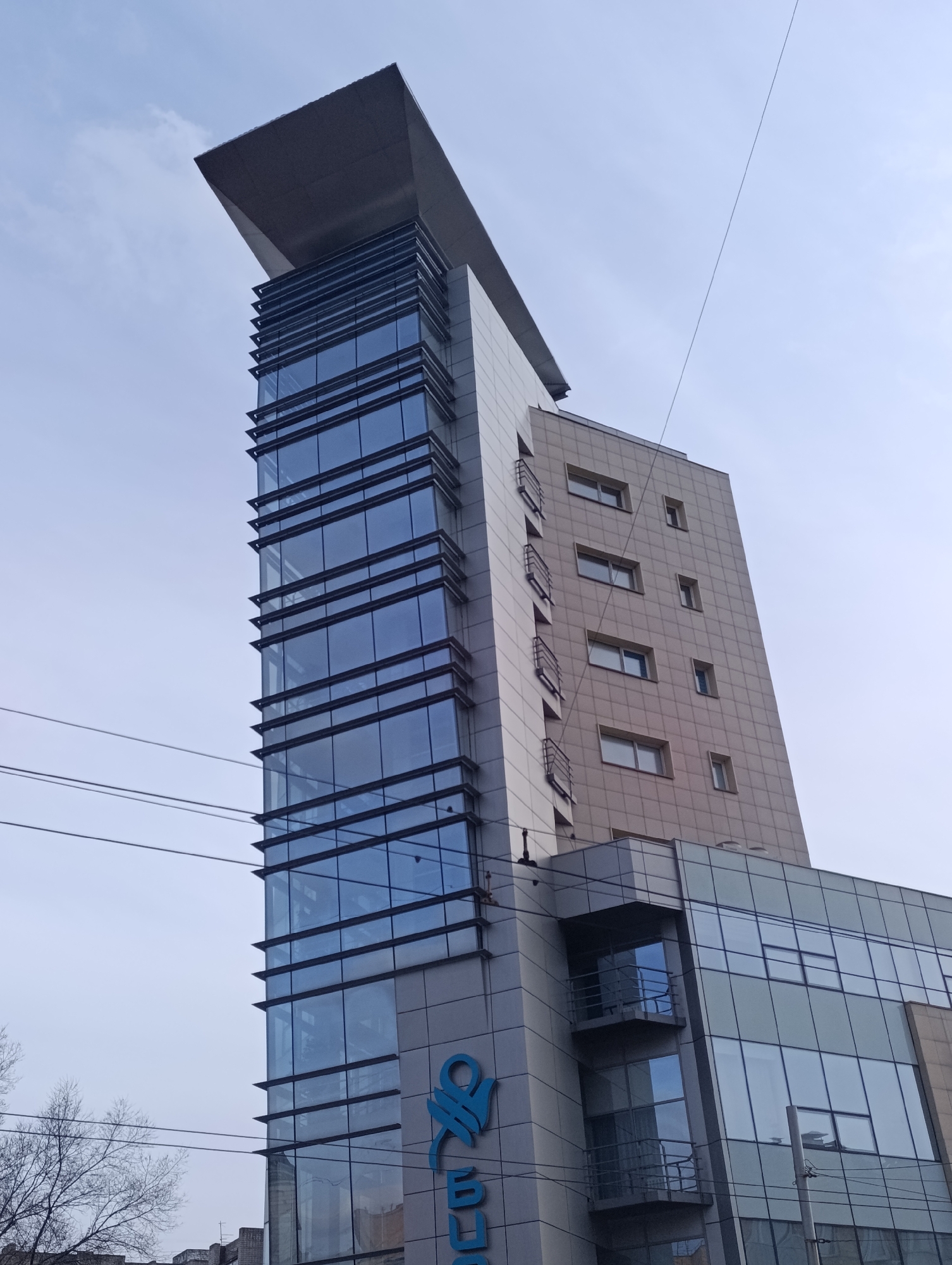 A low-angle shot shows a tall, modern building with a distinct architectural design in Krasnoyarsk, Russia. The building features a predominantly glass facade with horizontal dark trim, a cantilevered roof, and a tan brick-clad section. The building is located in a city, with power lines and the bare branches of trees in the foreground. The sky is overcast. A blue logo is visible on the lower portion of the glass facade.