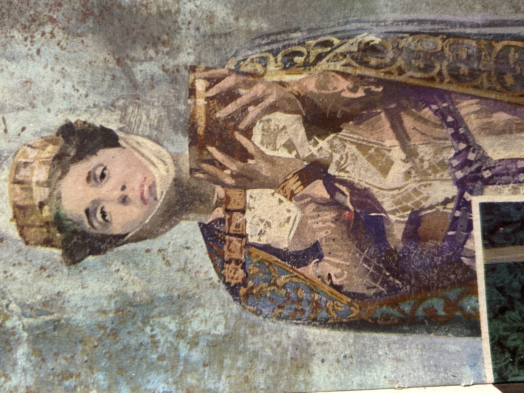 The image is a close-up, weathered, and somewhat faded artistic rendering of a person wearing a hat and ornate, layered clothing. The person's face is visible, with a somber expression, and a light-colored cloth is wrapped around their lower face. They are holding a rectangular object, possibly a book or a container, in their hands. The background is a textured, mottled surface in shades of blue, grey, and brown, suggesting an aged or distressed material. There is no discernible information within the image that indicates a specific location, such as the city of Santa Ana, Venezuela. The image is abstract and artistic in nature, lacking concrete geographical or contextual details.