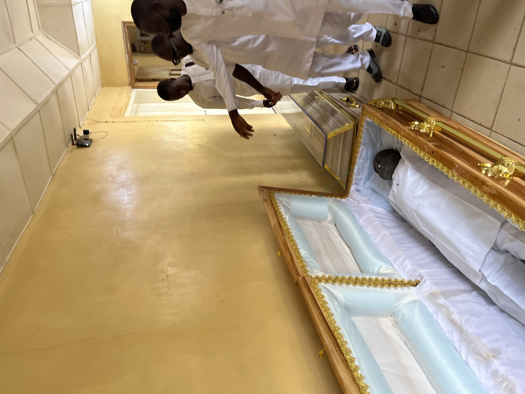 The image shows an interior scene, likely a building in Jos, Nigeria, given the apparent ethnic background of the individuals present. The room has yellow walls and a painted concrete floor. Three men dressed in white outfits and dark shoes or sandals are standing near two open coffins. One coffin, with a clear lid, appears to contain a body. The second coffin, finished with light wood trim and a light blue interior, is open and empty. A wireless router is visible attached to the wall.