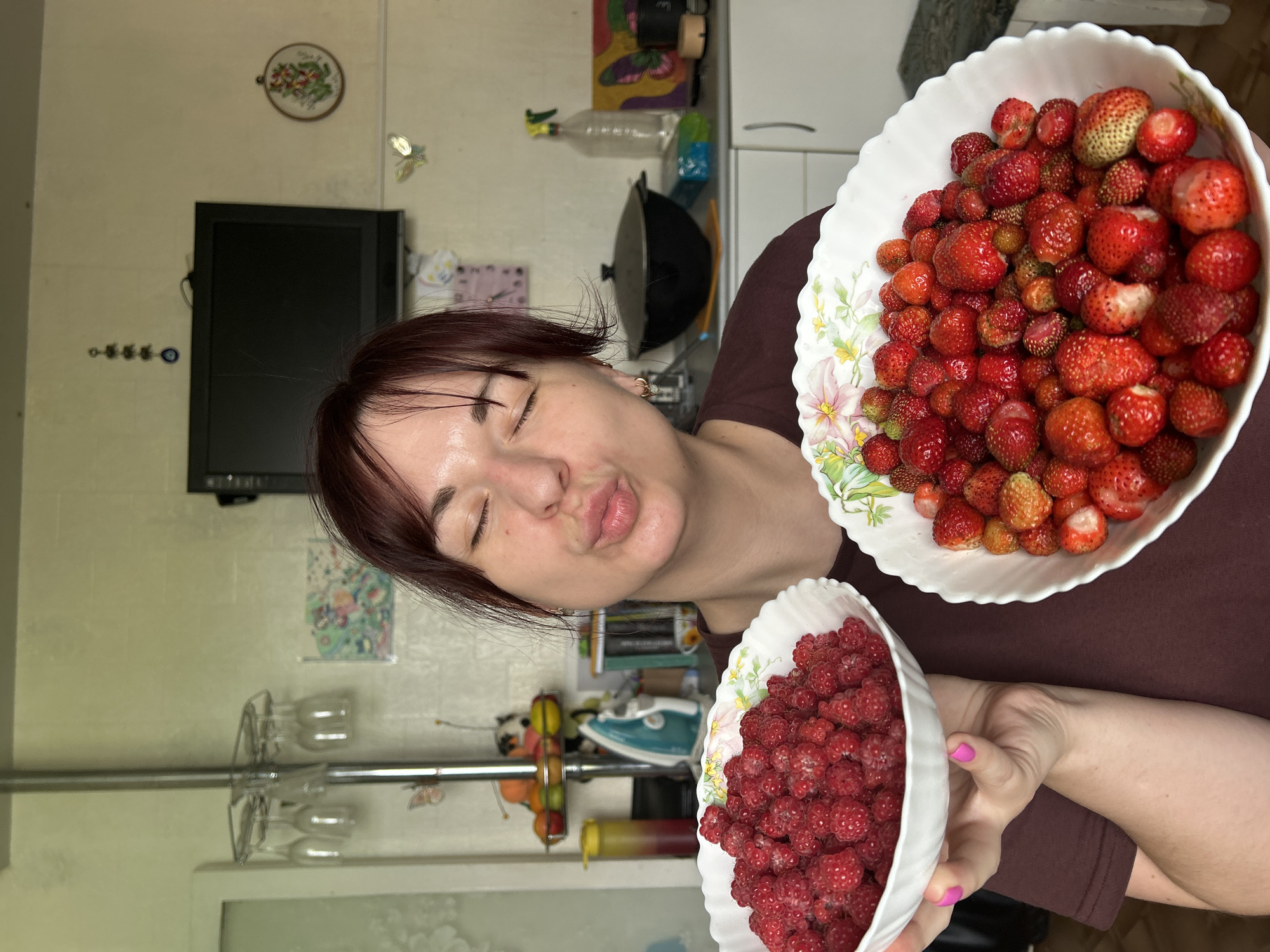 A woman with auburn hair and closed eyes puckers her lips as she holds two bowls of berries in front of her. The bowl on her left contains a large quantity of raspberries, while the bowl on her right is filled with strawberries. She is wearing a dark maroon t-shirt. The background appears to be a kitchen, with cabinets, a stovetop, and a microwave visible. There are also various decorative items on the walls and shelves, including glassware, a framed picture, and a hanging charm. The overall lighting is bright and natural. The specific location of Фоки, Russia is not evident in the image.