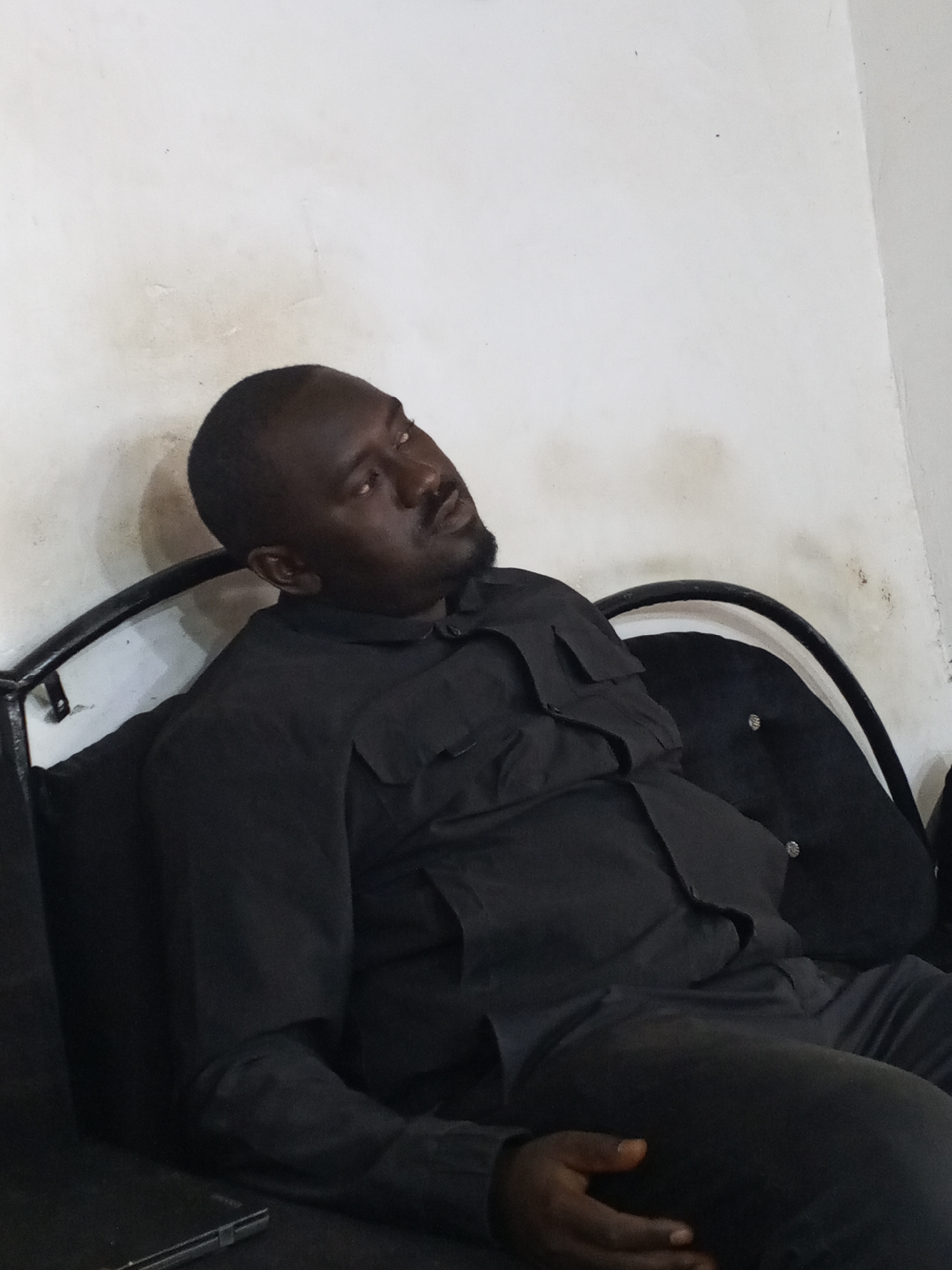 A man of African descent is seated on a black couch with a metal frame against a white wall. He is wearing a dark collared shirt with visible pockets and dark pants. His head is tilted back and to the right. His facial expression appears pensive. Part of a black mobile device is visible on the couch to the left. The location is likely indoors, within a residential or commercial setting in Gwong, Nigeria, based on the instructions.