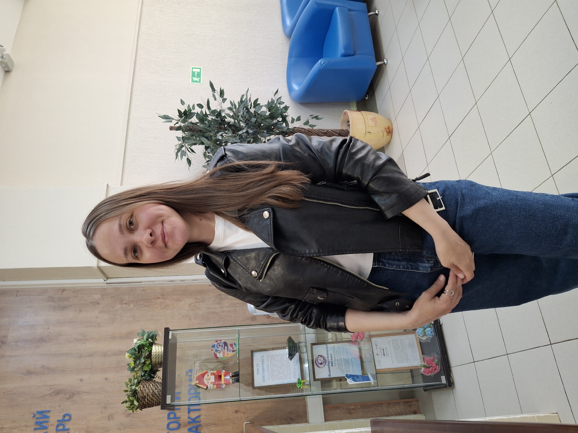 A woman stands indoors near a display case. She is wearing jeans and a black leather jacket. Behind her are blue chairs and a potted plant. The display case contains documents and decorative items. The location is in the city of Chaikovsky, Russia, as indicated by the inscription on a nearby wall.