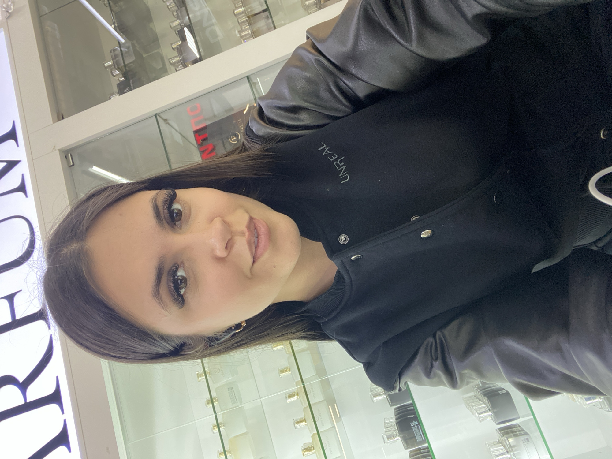 A young woman is depicted in a close-up portrait, oriented sideways with her head tilted towards the viewer, offering a slight smile. She has long, dark hair, dark eyes with pronounced eyelashes, and dark eyebrows. Her lips are a light, natural shade. She is dressed in a black jacket featuring dark, seemingly leather sleeves and a matte fabric body, with the word "UNREAL" visible in light-colored text on the left chest. Silver snap buttons are present on the jacket's front.
The background indicates an indoor commercial setting, likely a retail store in Chaikovsky, Russia. Several brightly lit glass display cases are visible, containing numerous small, clear and frosted bottles, suggesting the sale of perfumes or cosmetics. To the left, a large, illuminated sign displays partial white text, "ARBO", while another display case shows faint red text, possibly "ON", on a label. The lighting is bright and uniform, typical of an indoor retail environment. No specific activity is taking place beyond the woman posing for the photograph.