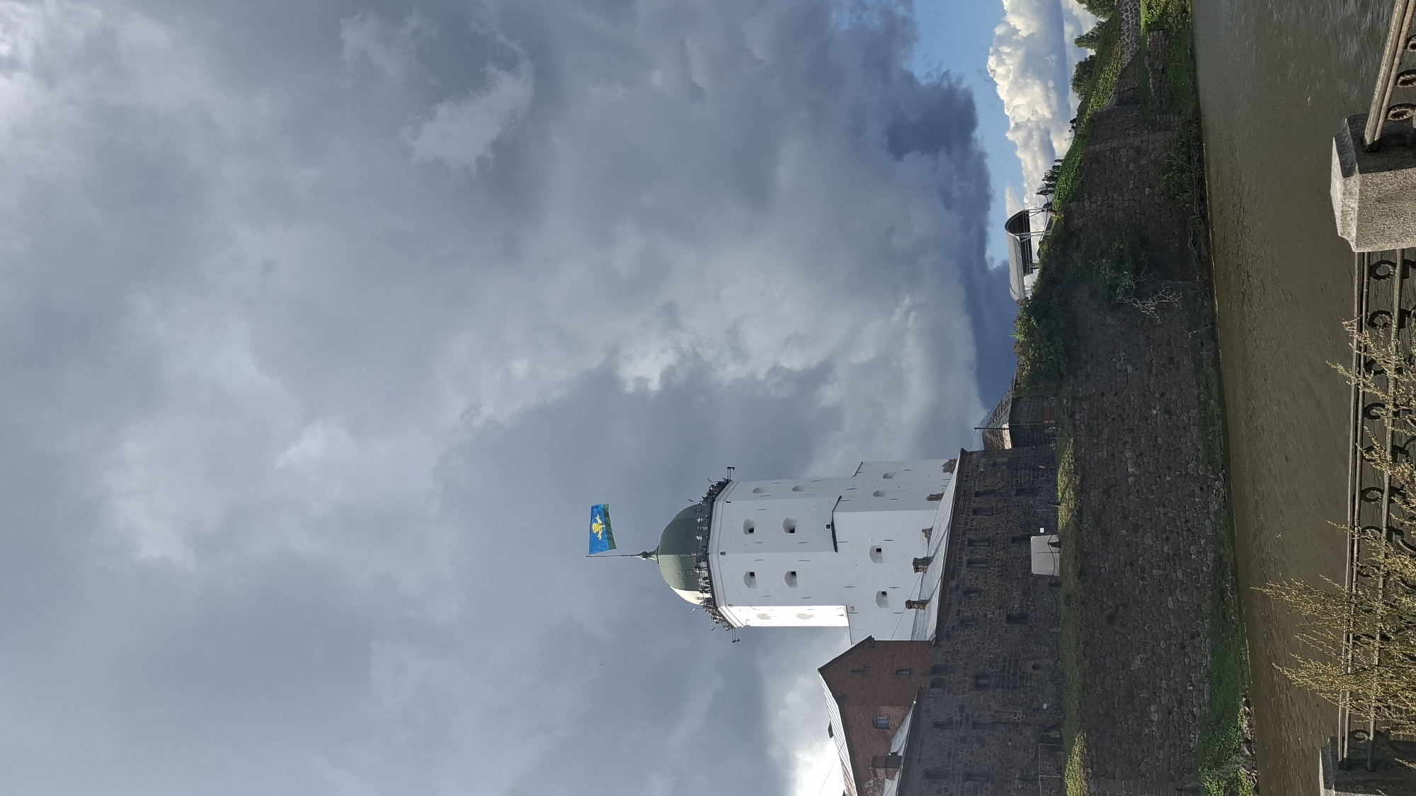 This is a vertical, eye-level shot of the Vyborg Castle in Vyborg, Russia, under a dramatic, cloudy sky. The castle is predominantly white with a green-domed tower, topped by a blue and yellow flag. A portion of a red brick building and a stone wall are visible to the left of the tower. The castle complex sits on a hill overlooking a body of dark water. In the foreground, a decorative metal fence with ornate posts and some dry grass peeking through it runs horizontally across the bottom of the frame. The sky is a mix of dark, heavy clouds and patches of bright blue with white cumulus clouds. Several small figures, presumably tourists, are visible on the castle grounds near the top of the hill to the right of the tower. The scene captures a historic landmark under dynamic weather conditions.