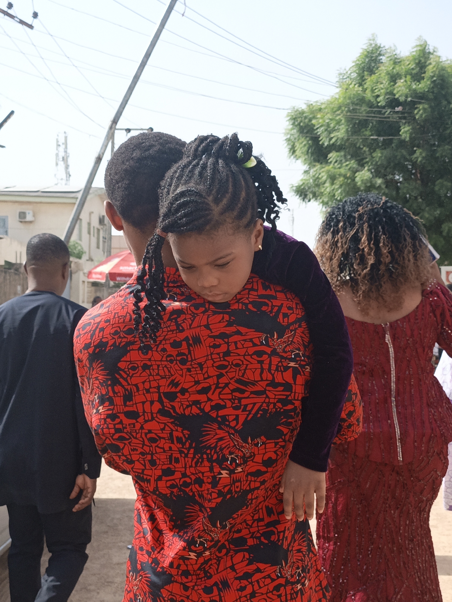 In Sabon Gari West, Nigeria, a man carries a young girl on his back. The girl wears a dark-colored velvet top and has her hair braided. The man wears a red shirt with a black pattern. The woman to their right wears a red sequined dress and has curly brown hair. Behind the man to the left, a man in a black suit walks away. Buildings and a tree are visible in the background.