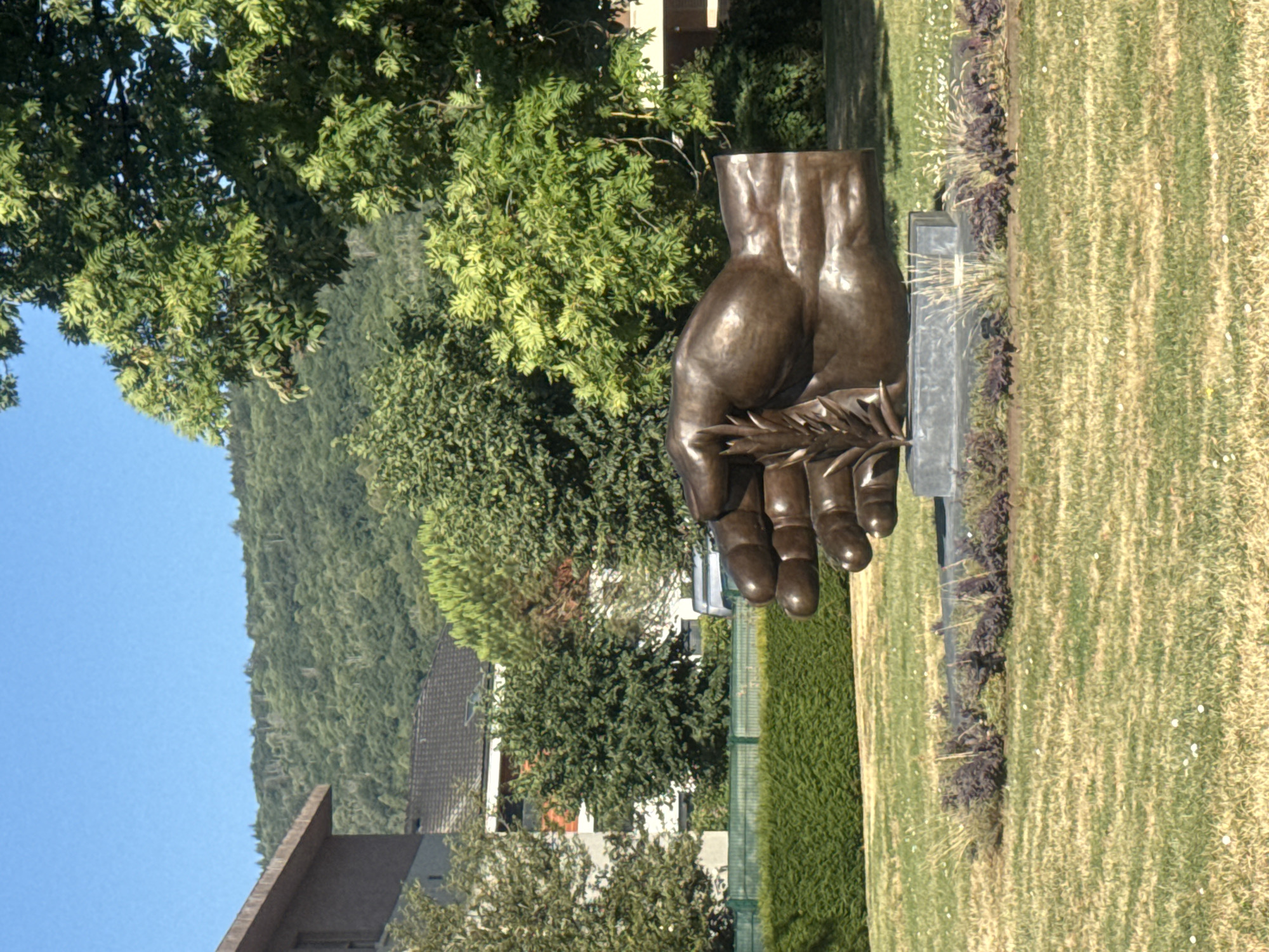 The image depicts a bronze sculpture of a cupped hand holding a single stalk of wheat, positioned outdoors on a sunny day. The sculpture is the central focus, situated on a grey stone base surrounded by low-lying, dark purple foliage and ornamental grasses. Behind the sculpture, a neatly trimmed green hedge forms a boundary, and beyond that, the facade of a modern building with a brown roof is visible. Further in the background, trees with lush green foliage and a distant, tree-covered hillside under a clear blue sky complete the scene. This sculpture is located in Divonne-les-Bains, France, specifically in the Jardin des Thermes.