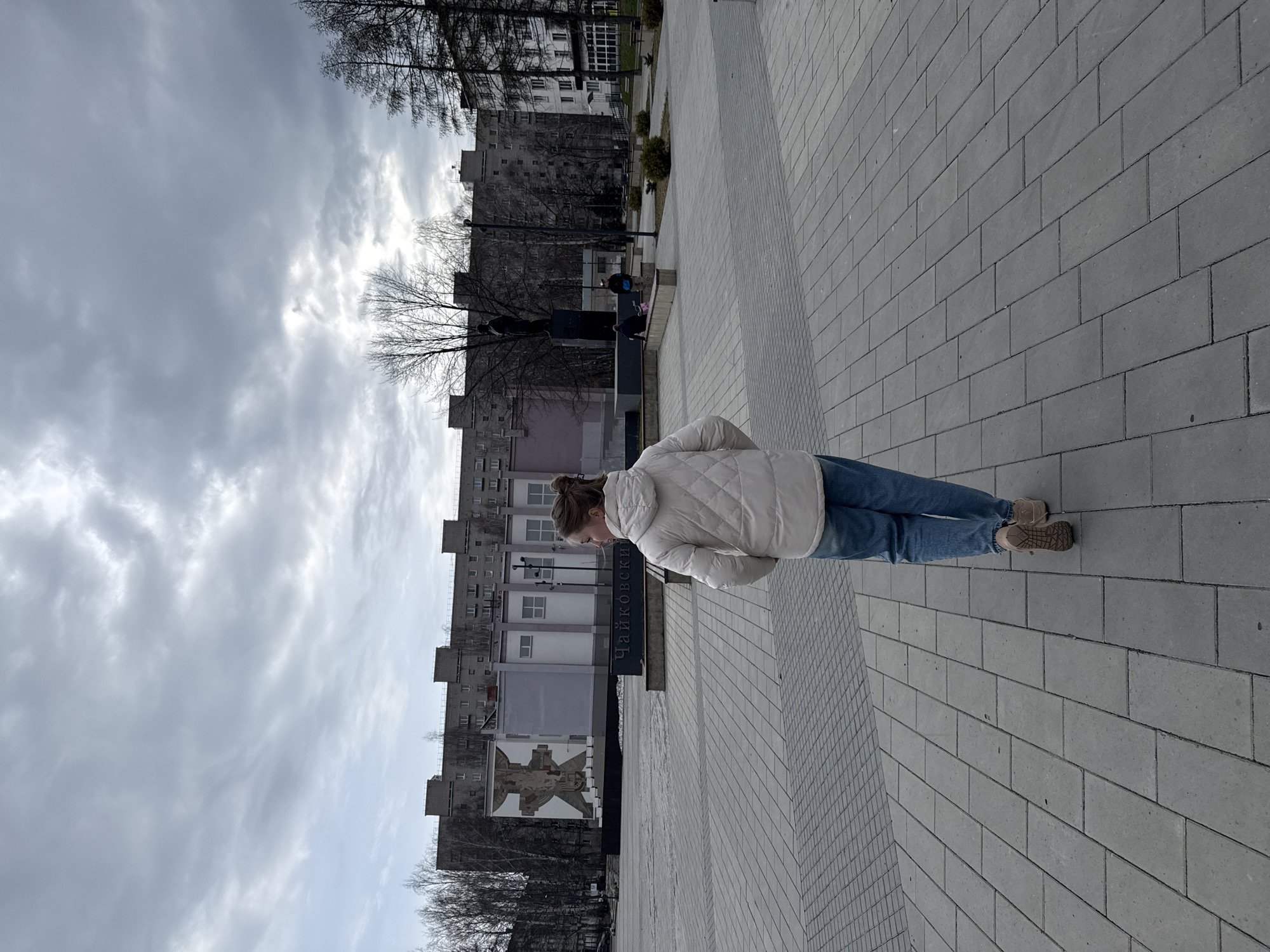 The image shows a woman standing on a paved plaza in the city of Unknown, Unknown. She is wearing jeans and a light-colored quilted jacket, with her head bent down as if looking at something. Behind her are Soviet-era apartment buildings and a monument. The sky is overcast. The photograph appears to be taken at street level, from a normal perspective.