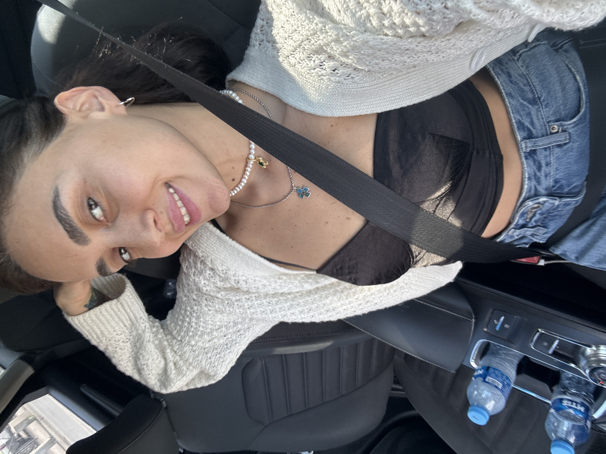 A woman is captured in a selfie from a high angle, seated in a car. She has dark hair, pulled back, and a light-colored, open-knit cardigan over a black bra. She is wearing blue jeans. A seatbelt is prominently visible across her chest. She is smiling and looking towards the camera. A delicate pearl necklace with a small pendant and a bracelet adorn her neck and wrist.
The setting appears to be the interior of a car, with the black leather seats and dashboard visible. In the background, two plastic water bottles with blue caps are placed in cup holders. The overall lighting suggests it is daytime, possibly sunny, given the highlights on the woman's face and clothing.
The image is oriented sideways. The woman's expression is happy and relaxed. There are no other people visible in the image.
There is some text visible on the water bottles, but it is not clearly legible. The location context is Tbilisi, Georgia.