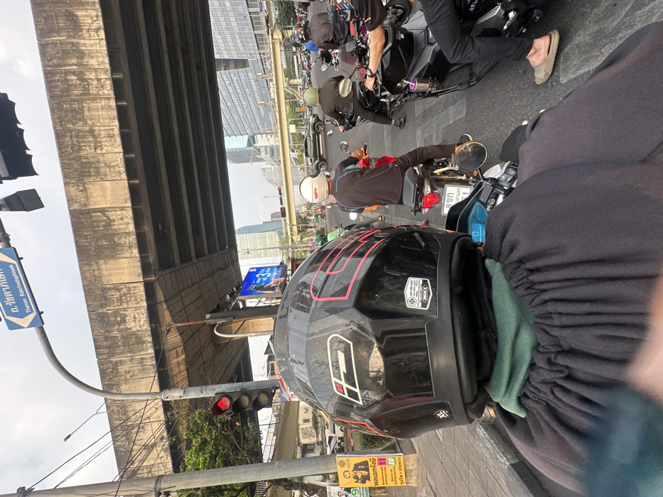 The image shows a person on a motorcycle, wearing a black helmet with red accents, waiting at a red traffic light under a concrete overpass. Other motorcycles and vehicles are also stopped. A blue street sign reading "Thanon Ratchadaphisek" (Ratchadaphisek Road) is visible. A nearby poster displays election information, mentioning "General Rangsi" and an anti-corruption message. The INDEX brand is visible on the helmet.