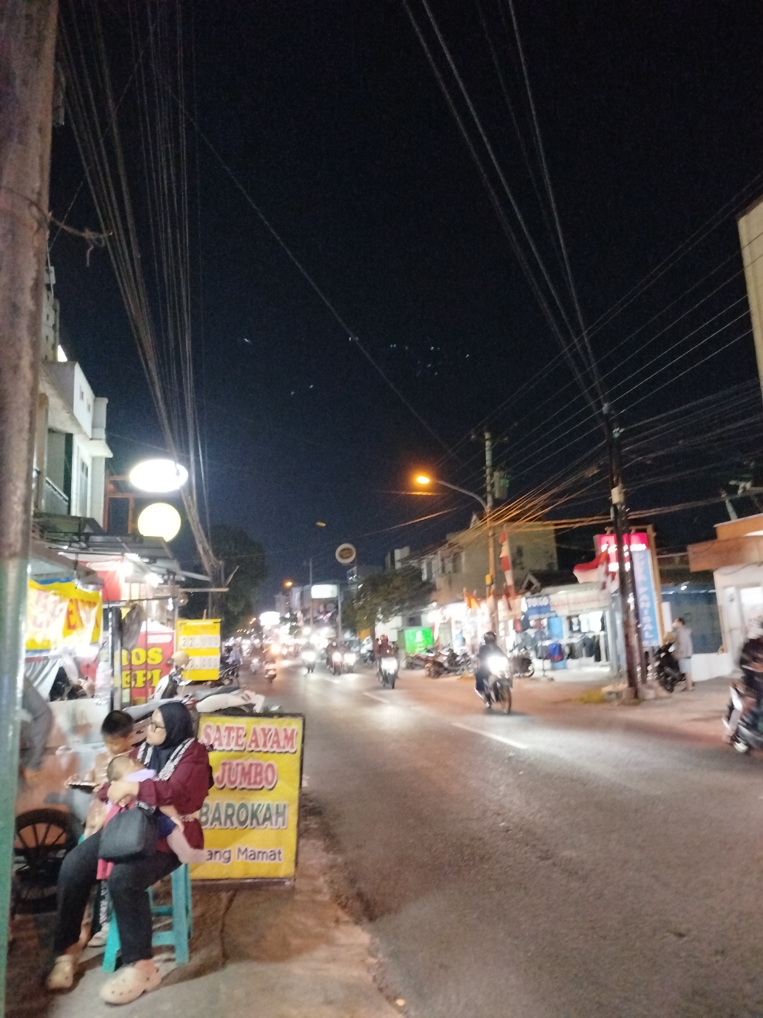 The image depicts a street scene at night in Surakarta, Indonesia. A woman wearing a hijab and a maroon jacket is seated on a light blue stool, holding a baby. To her right, a yellow sign with red text reads "SATE AYAM JUMBO BAROKAH" and below that "ang Mamam". The street is lined with shops and stalls, some of which are illuminated by bright lights. Multiple motorcycles are present, with some people riding them and others parked. Overhead, a complex network of power lines is visible against the dark sky. The overall atmosphere suggests a bustling urban environment at night, likely a commercial area with food vendors.