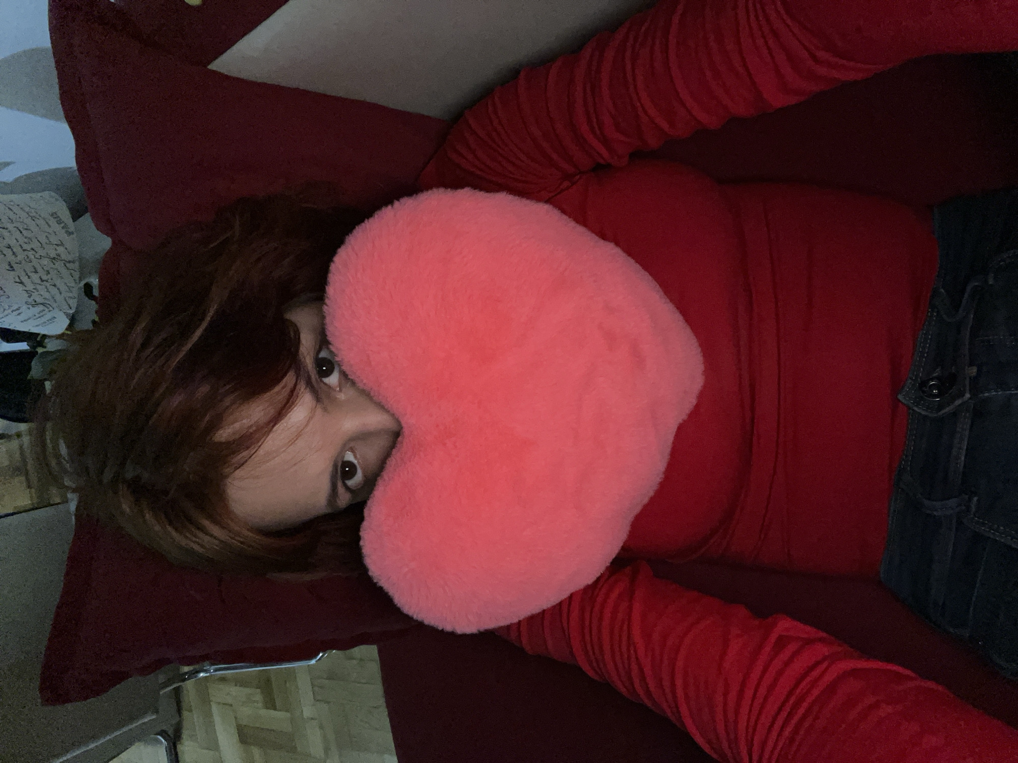 A woman with brown hair is lying on a red surface, possibly a bed or couch, with a pink heart-shaped pillow covering part of her face. She is wearing a red long-sleeved shirt and dark denim jeans. The setting appears to be indoors, with a partial view of a patterned rug and other items in the background. The image provides no data about the city of Boreti, Montenegro.