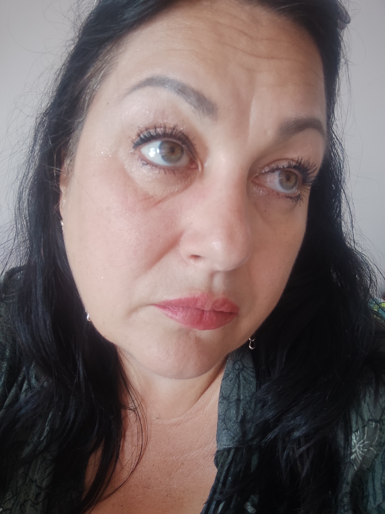 This is a close-up portrait of a middle-aged woman with dark hair and light brown eyes. She is looking off to the side with a slightly worried or contemplative expression. Her makeup includes mascara, eyeliner, and lipstick. She is wearing a dark green top with a patterned design. Small hoop earrings are visible. The lighting suggests an indoor setting with natural light coming from the side. The background is a plain, light-colored wall. There are no visible signs of the location in Sochi, Russia.