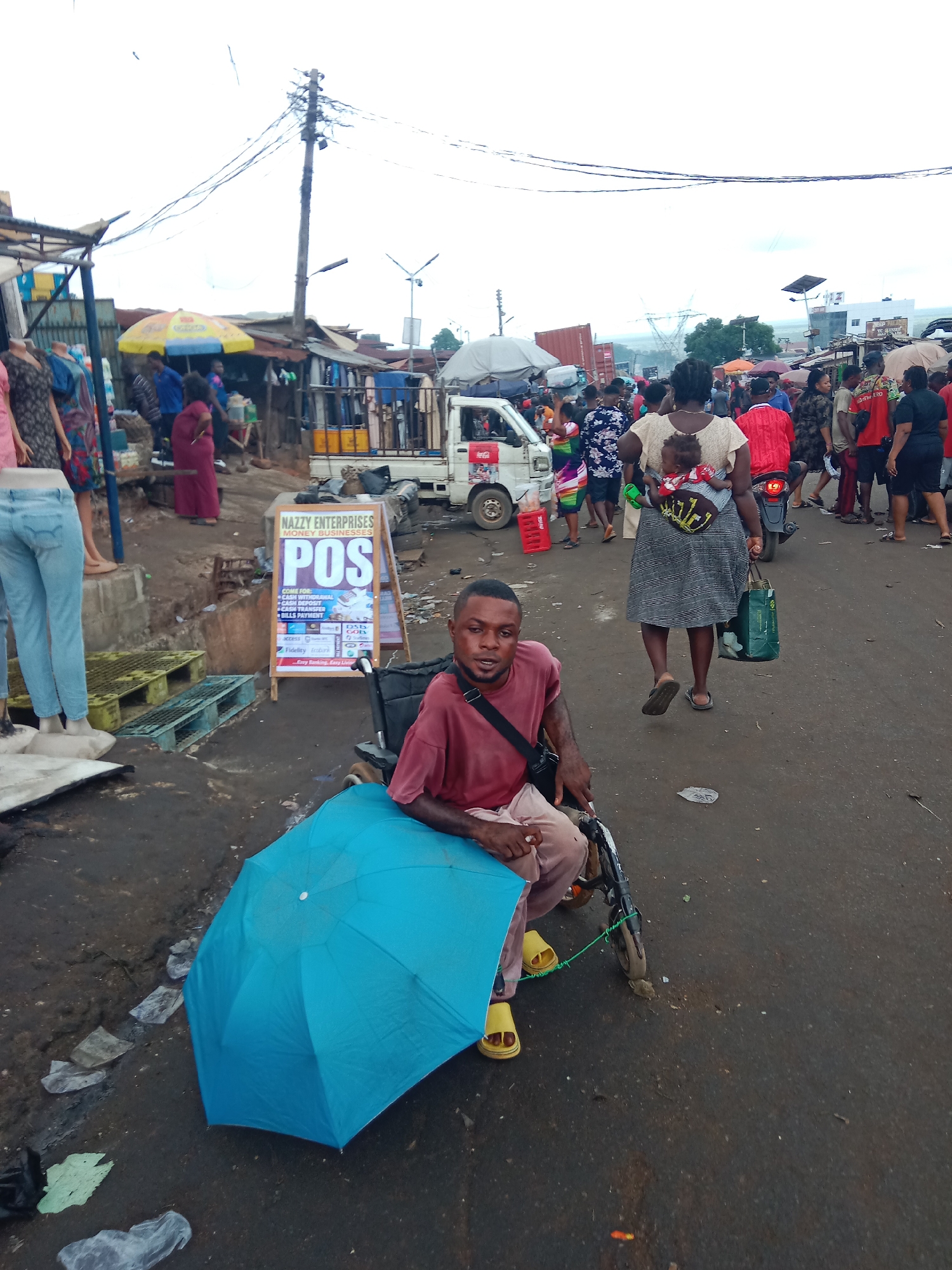 The image captures a crowded street scene in Obosi, Nigeria. A man in a wheelchair sits to the side of the road, under a blue umbrella. He is wearing a reddish-brown shirt and light-colored trousers with yellow sandals. A POS sign is visible next to him, advertising money transfer services. In the background, many people are walking along the street. A woman carries a baby on her back, and vehicles are present in the background. The ground appears wet and littered. Overhead, power lines crisscross the sky.