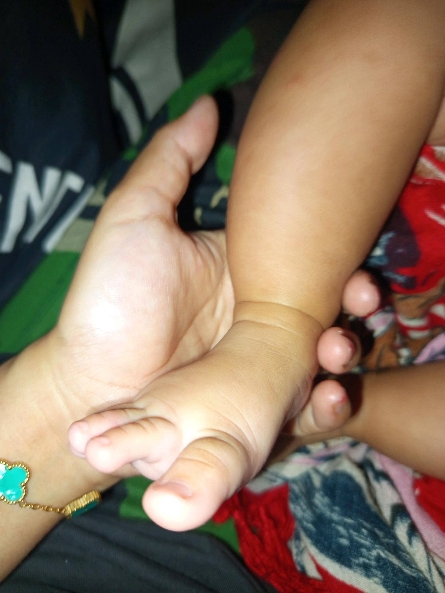 An adult hand holds a baby's foot. The baby's foot is positioned with the toes pointing upwards, and the heel is resting against the adult's palm. The baby's toes are curled slightly. A portion of the baby's leg is visible above the foot. The adult is wearing a bracelet on their wrist. The background consists of a dark fabric with a pattern, and what appears to be a colorful blanket. The location is Salebu, Indonesia.