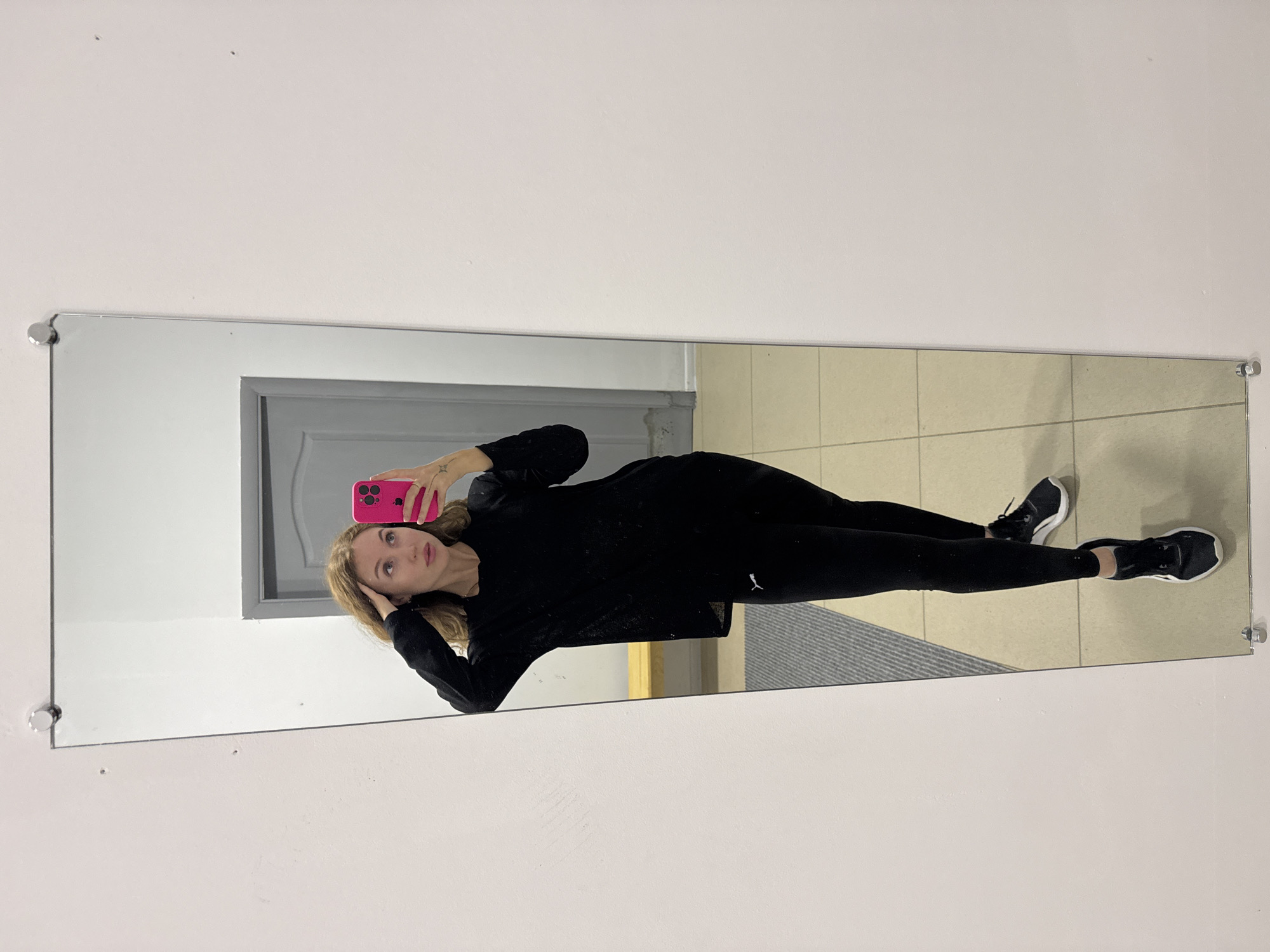 A young woman is taking a mirror selfie in an indoor setting. She stands in front of a large, rectangular mirror mounted on a plain white wall.
The woman has light-colored hair and is wearing an all-black athletic ensemble, including a long-sleeved top and leggings. A white Puma logo, depicting a leaping feline, is visible on the upper left thigh of her leggings. She completes her outfit with black athletic shoes that have white accents.
She holds a bright pink smartphone in her right hand, positioned to capture her reflection. Her left hand is casually placed near her head, and she looks directly into the phone's camera. A small, star-like tattoo is visible on the back of her right hand.
The reflection in the mirror shows a tiled floor made of light-colored square tiles and a dark, textured rug or mat under her feet. In the background of the reflection, a grey door is partially visible. The overall environment appears to be a functional indoor space, possibly a hallway or changing area in a gym or public building. The lighting is consistent with an indoor setting, without strong natural light. The location context for this image is Chaikovsky, Russia.