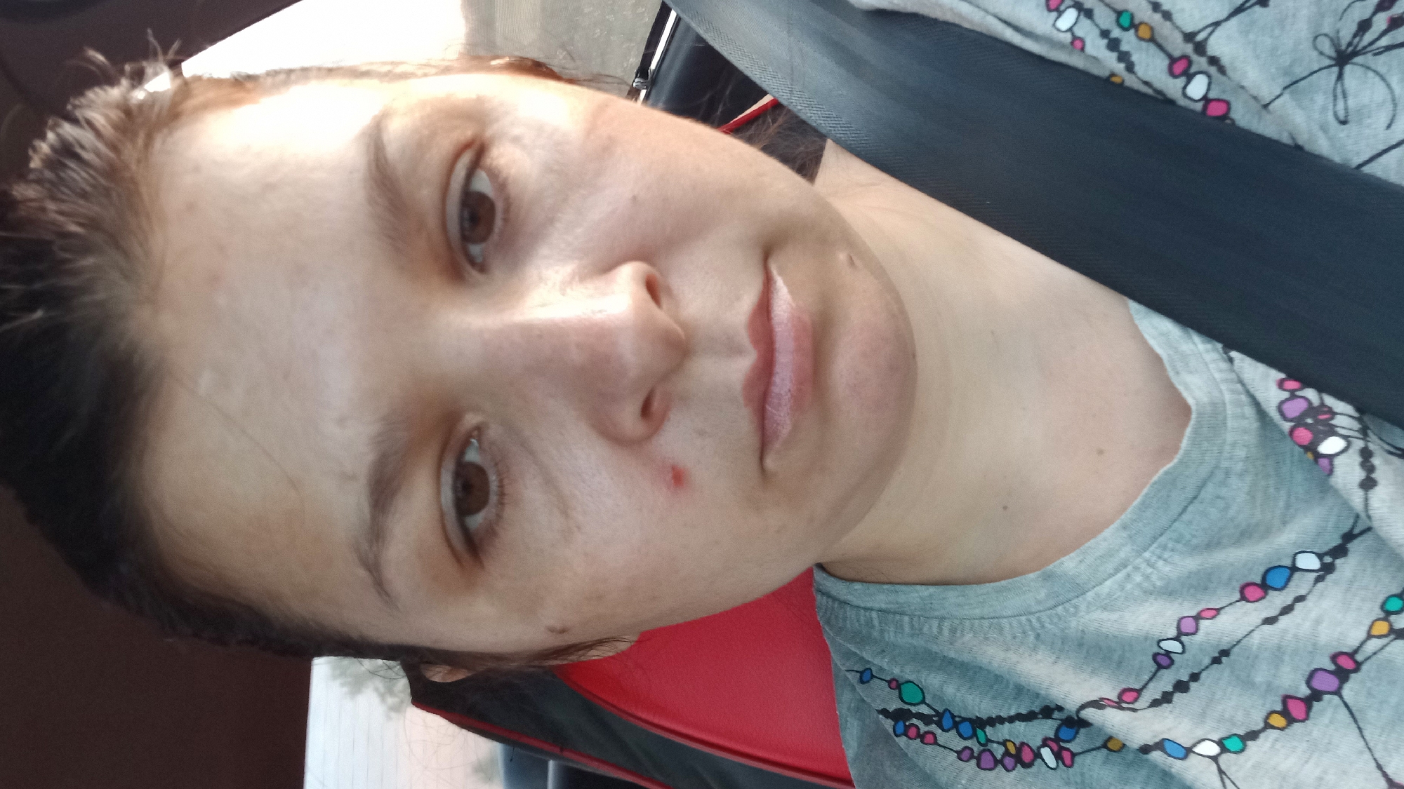 A close-up, head-and-shoulders selfie shows a woman with brown hair pulled back and brown eyes. She is wearing a grey t-shirt with a colorful beaded necklace pattern. A red seatbelt is visible across her chest. She has a red blemish on her left cheek and a mole on her chin. The background appears to be the interior of a car, with a red seat and a dark dashboard. The image is taken in Klyntsi, Ukraine.