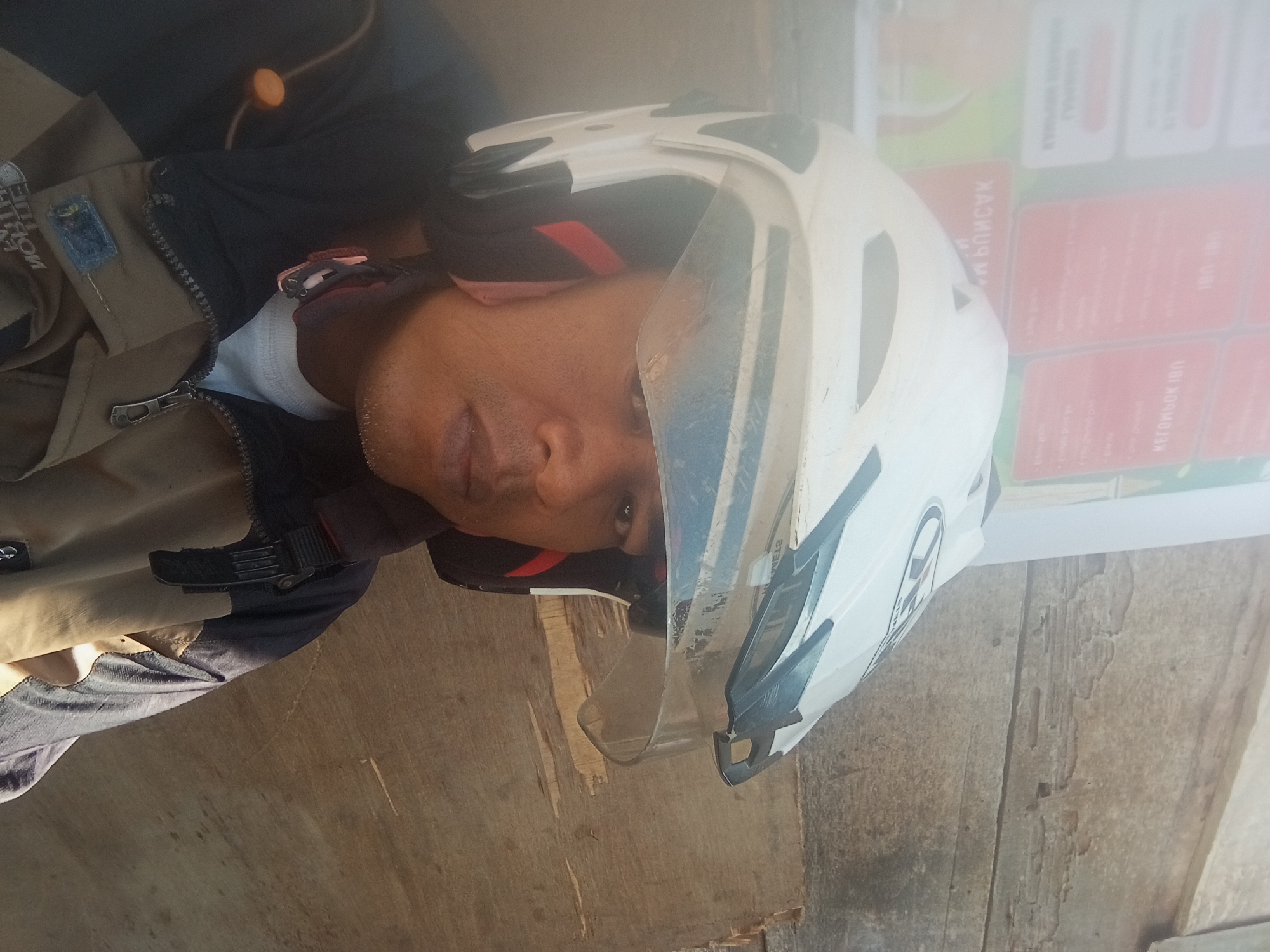 A man wearing a white and black helmet and a tan and dark blue jacket is pictured in front of a weathered wooden surface. The man's face is partially visible, and he appears to be looking towards the camera. A blurred background shows red and white signs with text. The setting is outdoors, likely on a street in Pagar Alam, Indonesia. The overall lighting suggests it is daytime.