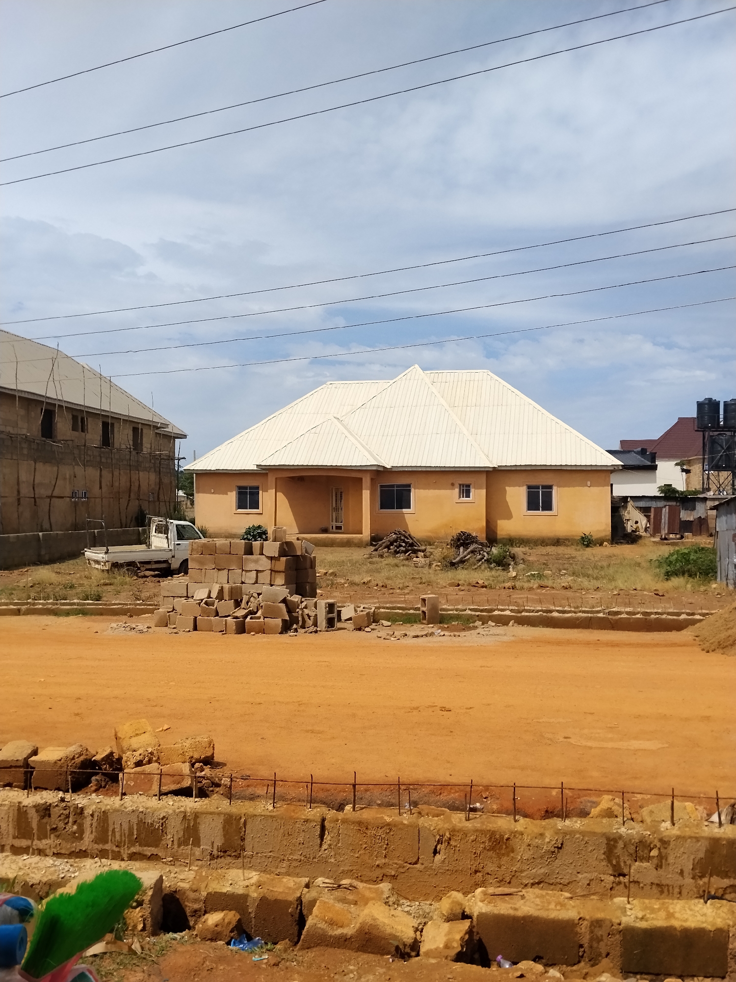 In Laminga, Nigeria, a construction site is depicted with a single-story, tan-colored house featuring a light-colored metal roof. Adjacent to this house, another building, possibly under construction, stands with scaffolding. The foreground is dominated by a dirt road and a pile of cinder blocks. A white pickup truck is parked near the cinder blocks and the house. Power lines crisscross the sky, and the environment appears dry and dusty. There are no people visible in the image.