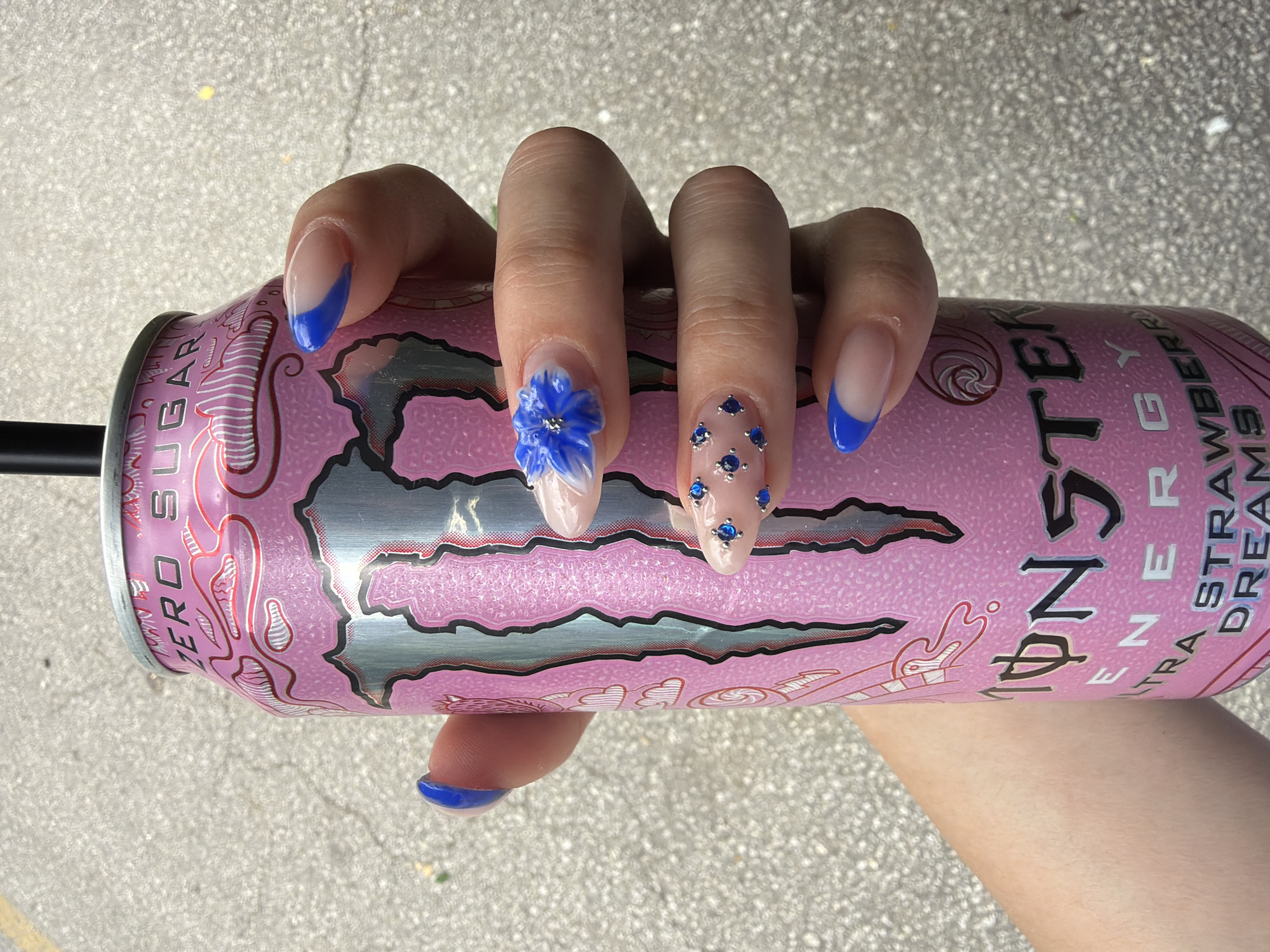 A human hand holds a pink can of Monster Energy Ultra Strawberry Dreams drink. The fingernails of the hand are manicured with a clear base and blue tips, with one nail featuring a 3D blue flower and another adorned with several small blue gemstones. The can is positioned against a background of light-colored pavement. The location is not discernable from the image, thus the city of Pazardzhik, Bulgaria cannot be confirmed.