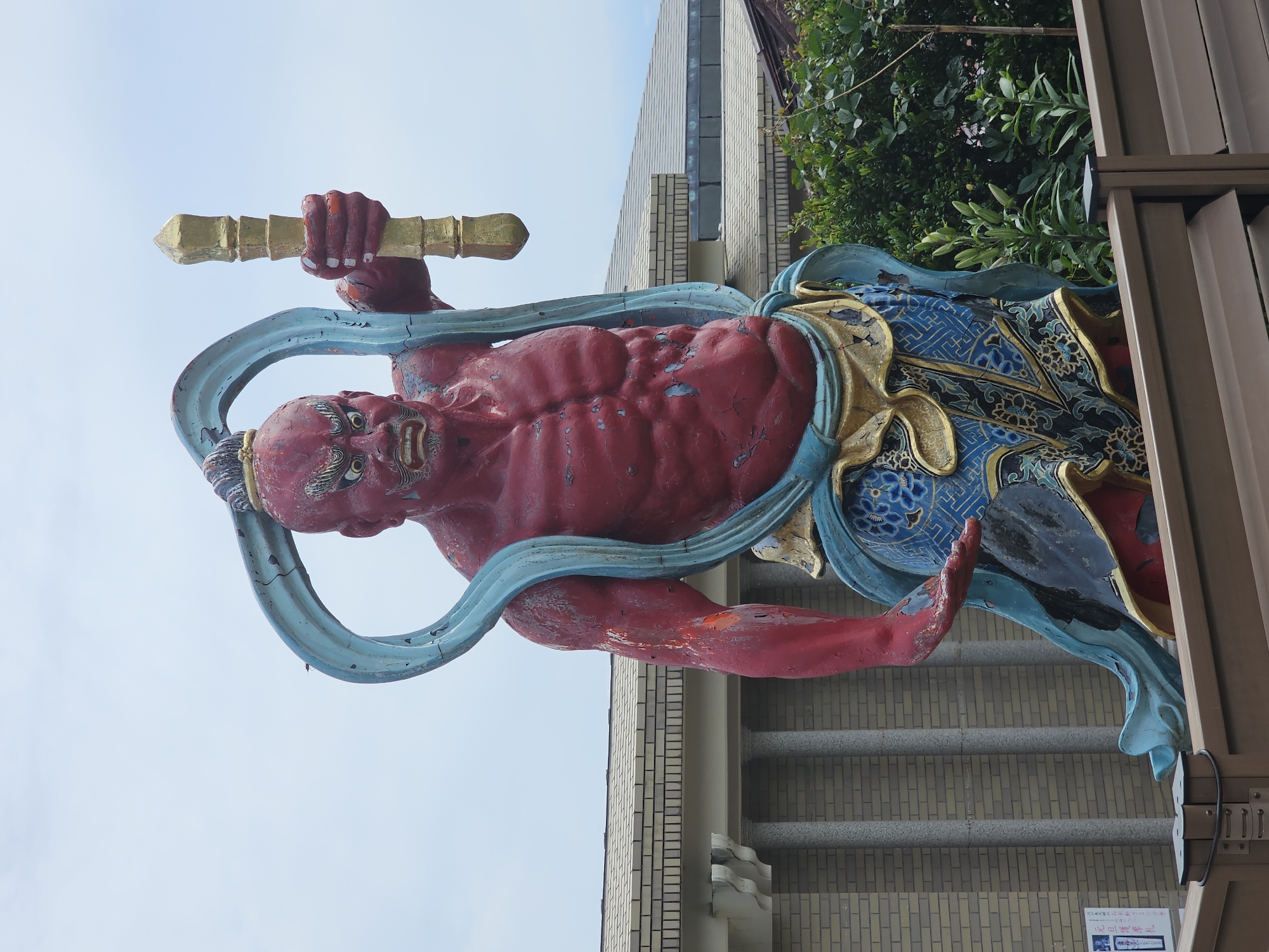 A full shot captures a vibrant, weathered statue of a red-skinned, muscular deity. The deity is depicted with a fierce expression, bulging eyes, and an angry grimace. A blue sash or ribbon encircles its neck and flows down its body, contrasting with the statue's skin and its ornate blue and gold patterned lower garment. In its right hand, the deity holds a golden, staff-like object. The statue stands outdoors, with a building featuring a brick facade and classical columns visible in the background. Lush green foliage is also present. The sky above is overcast and pale blue. This statue is located in front of a building in the city of Fujisawa, Japan.