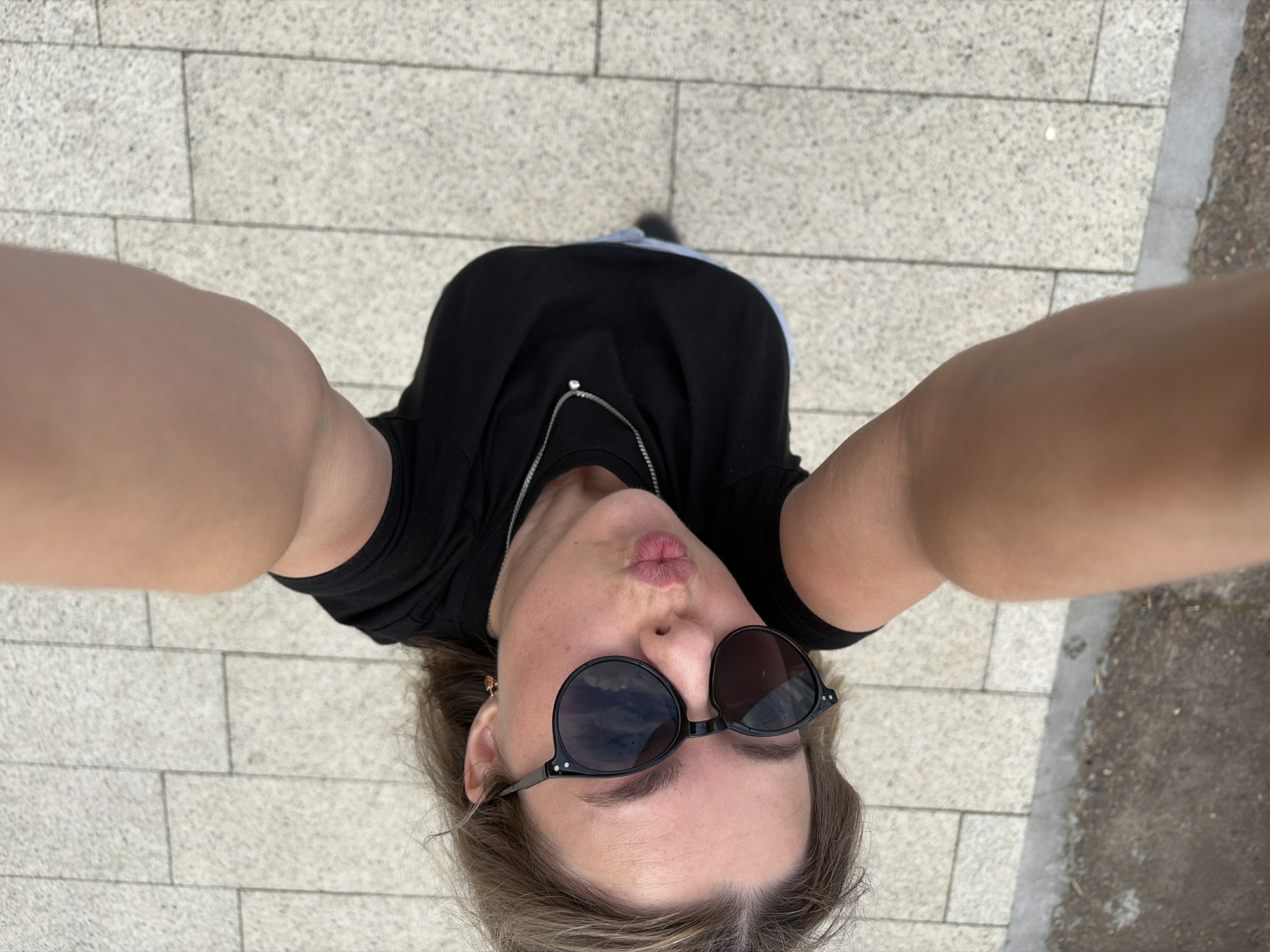 A high-angle, outdoor, full-body selfie is captured from the perspective of a person standing on a paved area composed of light-colored rectangular tiles. The individual is wearing a black t-shirt and light-colored pants, with sunglasses on their face and their lips pursed. Their arms are outstretched, holding the camera. The background consists of the tiled pavement and a darker, possibly concrete or asphalt edge. The lighting suggests it is daytime. The image does not contain any identifiable landmarks or features that would indicate a specific location within Chaikovsky, Russia.