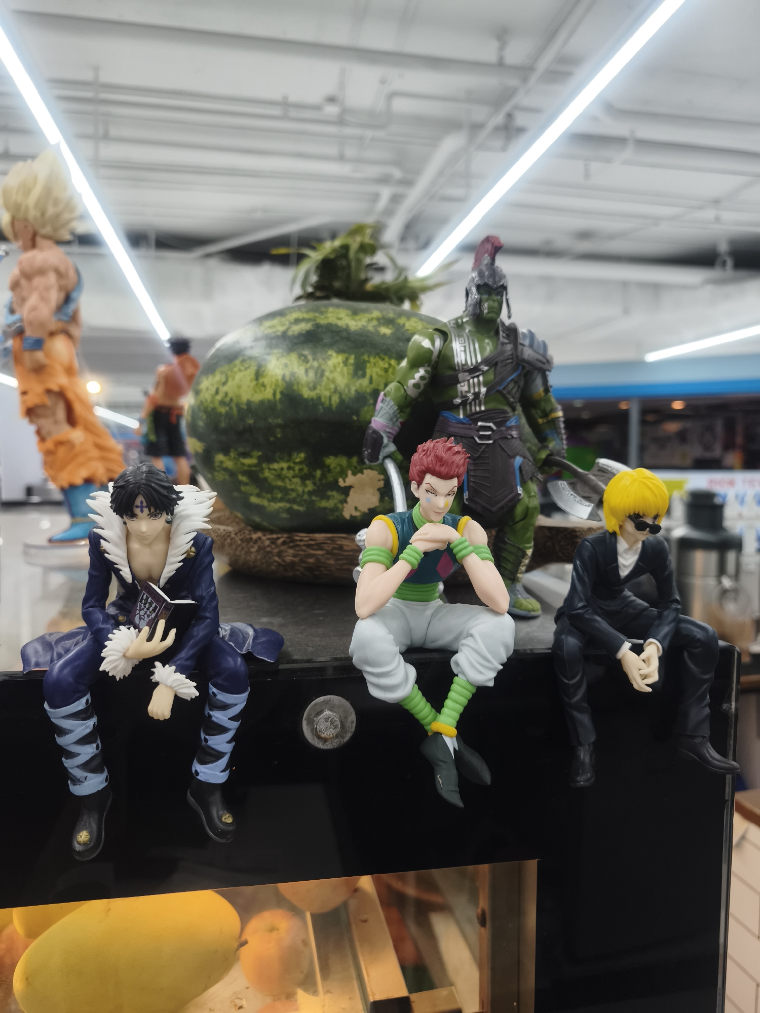 The image shows an assortment of figurines placed atop a black surface, likely a display case, possibly within a supermarket setting. The figurines are of various characters, including those from the anime "Hunter x Hunter," with Chrollo Lucilfer, Hisoka Morow, and an individual resembling Kurapika. Additional figurines include a green Gladiator Hulk-like figure with an axe, a watermelon, and a couple of Dragonball Z Goku figures. The environment appears to be a well-lit supermarket with fluorescent lights overhead. A fruit cooler is visible below the figures, containing mangos and oranges. The photo was taken in the city of บ้านบางรักษ์, Thailand.