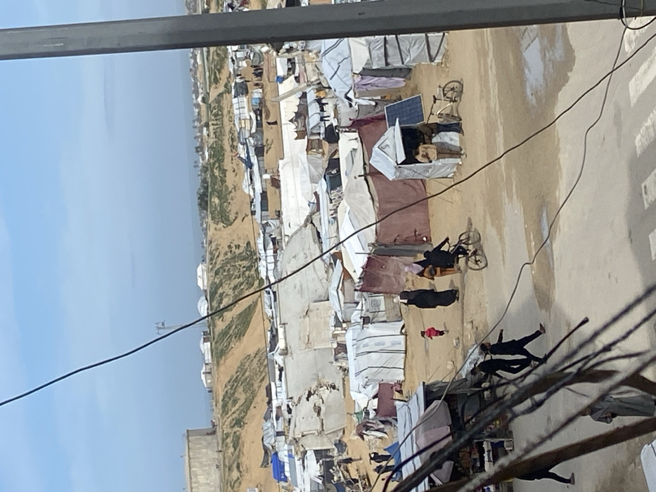 The image depicts a crowded, makeshift settlement, identified as being in Khan Yunis, Palestinian Territory, under a clear to partly cloudy blue sky.
In the background, a large, cylindrical, grey industrial tank stands atop a sandy, sparsely vegetated hill. Beyond this hill, the Mediterranean Sea is visible on the horizon, with what appears to be a coastal settlement or town along the shoreline. Scattered across the hilltop are several white dome-shaped structures and other temporary shelters.
The mid-ground is dominated by a dense array of temporary shelters and tents, constructed from various materials including white tarpaulins, striped fabrics, and maroon-colored sheets. Several structures show signs of damage or collapse, with torn and crumpled materials. Clothes are visible hanging on lines between some of the tents. The ground in this area is mostly dirt and sand, with visible wet patches or puddles.
Within the mid-ground, multiple individuals are present. A small child in a pink outfit runs on a dirt path. Nearby, three adults walk; two are wearing dark, long garments and head coverings, and one is in lighter attire. To their right, a person on a bicycle moves across the dirt path. Further right, two men stand near a small, white, stall-like structure that has a solar panel affixed to its side. A bicycle is parked next to this stall.
In the foreground, a paved section of ground is visible where several individuals are walking. One person wears a light-colored, long garment, while others are in dark clothing. Overhead, dark utility cables crisscross the frame, partially obscuring the view, originating from a vertical grey structure on the far right edge of the image.