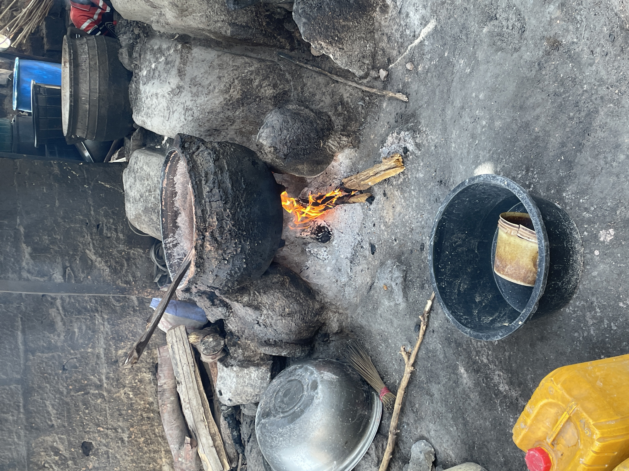 The image shows an outdoor cooking scene in Jos, Nigeria. A large, blackened pot sits atop a makeshift stove constructed from rocks and burning wood. To the right of the stove, there is a black plastic bucket containing a yellow container and a yellow plastic container sits on the ground. Other objects include a metal bowl, various containers and what appears to be a small broom. The environment consists of a rough, concrete surface and a stacked configuration of metallic containers and dark stone. A person is visible behind the metallic containers on the upper left of the photo.