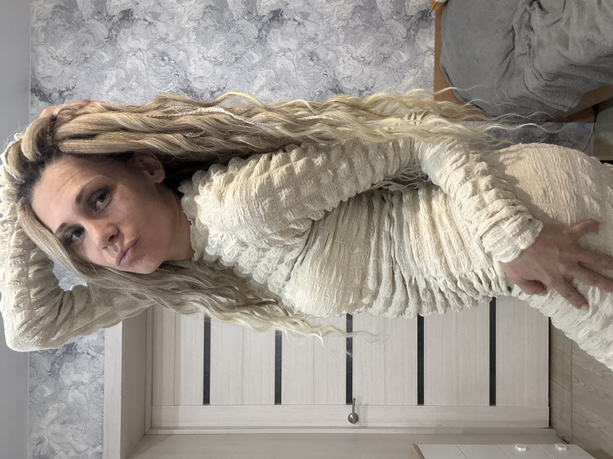 A woman with blonde dreadlocks is posing in a long-sleeved white dress. She is holding her hand up near her head and is also holding her hip with the other hand. She is indoors, with a white door with black accents behind her and a gray and white marble-patterned wall to her left. The top right corner of the image shows a gray fabric on top of a wooden surface. There is no information available that indicates that the photograph was taken in Шебунино, Russia.