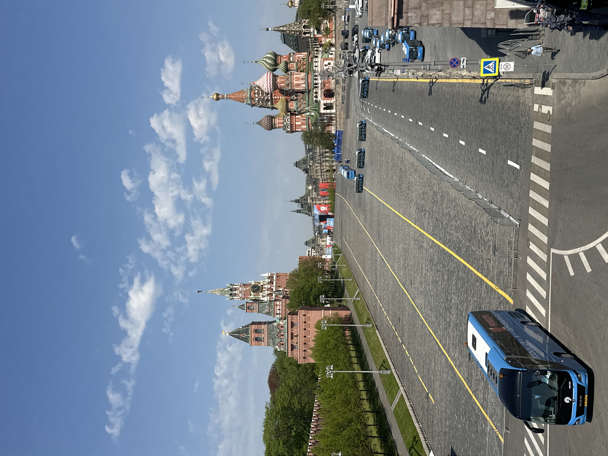 The image depicts a street scene in Moscow, Russia, featuring Red Square. St. Basil's Cathedral is visible in the background, characterized by its colorful and ornate domes. Adjacent to it, a portion of the Kremlin wall can be observed, along with the Kremlin towers. The street is paved with cobblestones and marked with yellow lines, indicating lanes. A blue and white bus is positioned on the right side of the image, near a pedestrian crossing. The sky is blue with scattered white clouds. The area appears to be prepared for an event, given the number of identical buses parked along the road.