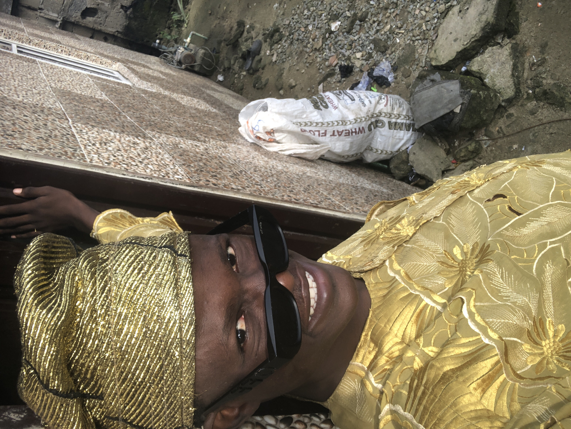 The image is a close-up, high-angle, outdoor shot of a young Black male in Ado, Nigeria, wearing a gold embroidered traditional outfit and a matching gold headwrap. He is also wearing black sunglasses. He is smiling broadly, showing his teeth, and looking directly at the camera. The background consists of a beige, textured building wall and a pile of gravel and debris. A white bag, possibly containing wheat flour, is leaning against the wall. A pump and some wires are also visible in the background, suggesting an outdoor or semi-outdoor location possibly related to agricultural or industrial activity. The man is positioned to the left of the frame, while the building and debris occupy the right side.
