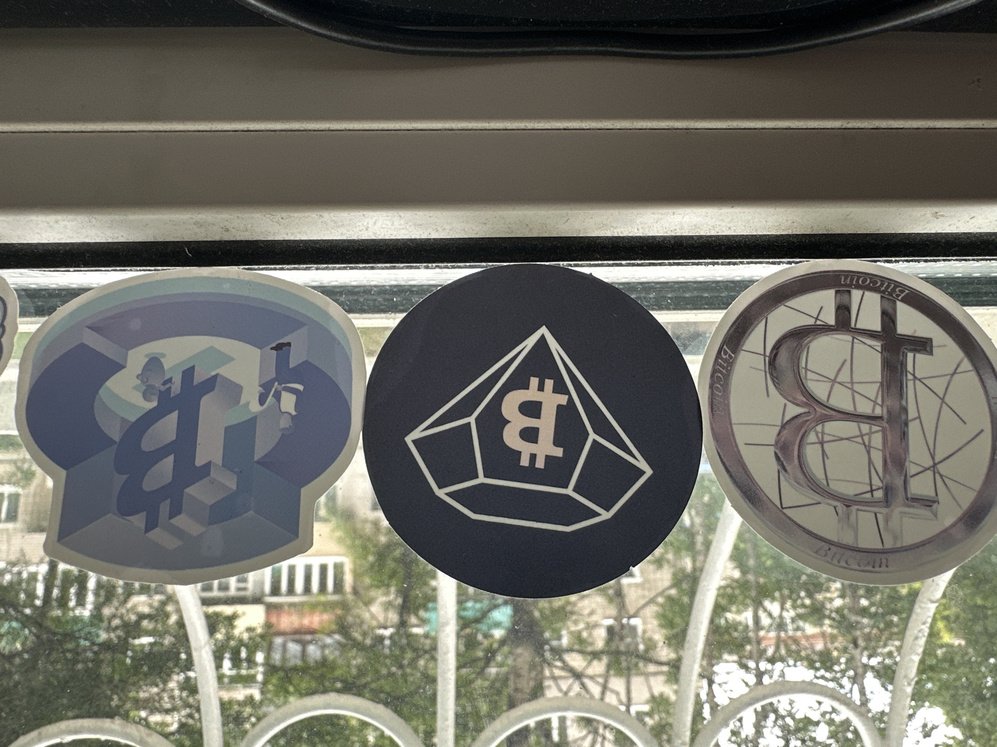 Three stickers are placed on a windowsill, with a window and foliage visible in the background. The stickers appear to be related to cryptocurrency. The leftmost sticker is circular and features a metallic-looking "B" symbol with lines radiating from it, and the word "Bitcoin" is visible at the bottom. The middle sticker is also circular, with a dark background and a white outline of a diamond shape containing a "B" symbol. The rightmost sticker is die-cut and depicts a stylized 3D "B" symbol within a geometric design, with small figures of people around it. The windowsill is white and shows some dust and grime. The environment outside the window suggests an urban setting with buildings and trees. The image was taken in Nakhodka, Russia.
