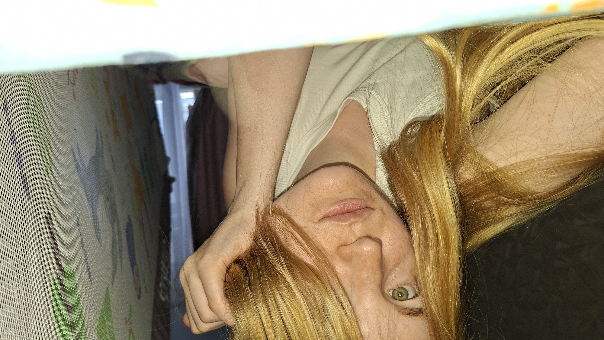 The image displays a close-up view of a woman, rotated 90 degrees clockwise from a standard upright orientation. She has long, reddish-blonde hair, fair skin, and visible freckles. Her face occupies the lower-right portion of the frame, with one green eye (her left eye, if oriented upright) looking directly towards the viewer. Her lips are slightly parted, and her expression appears neutral, possibly gentle or observant. She is wearing a light-colored t-shirt. Her right arm is bent, with her hand resting near her head, suggesting she is leaning over something.
The perspective is from below, looking upwards, as if the camera is placed inside an enclosure. To the upper-left of the frame, a textured mesh barrier, indicative of a playpen or crib, is clearly visible. Behind this mesh, a colorful mat with various cartoon-like illustrations, including animal shapes, is present, suggesting a child's play area. In the background, natural daylight streams through a window, indicating it is daytime. A dark curtain or fabric is also visible in the distance. No discernible text is present in the image. The scene is indoors, within a domestic setting in Chaikovsky, Russia.