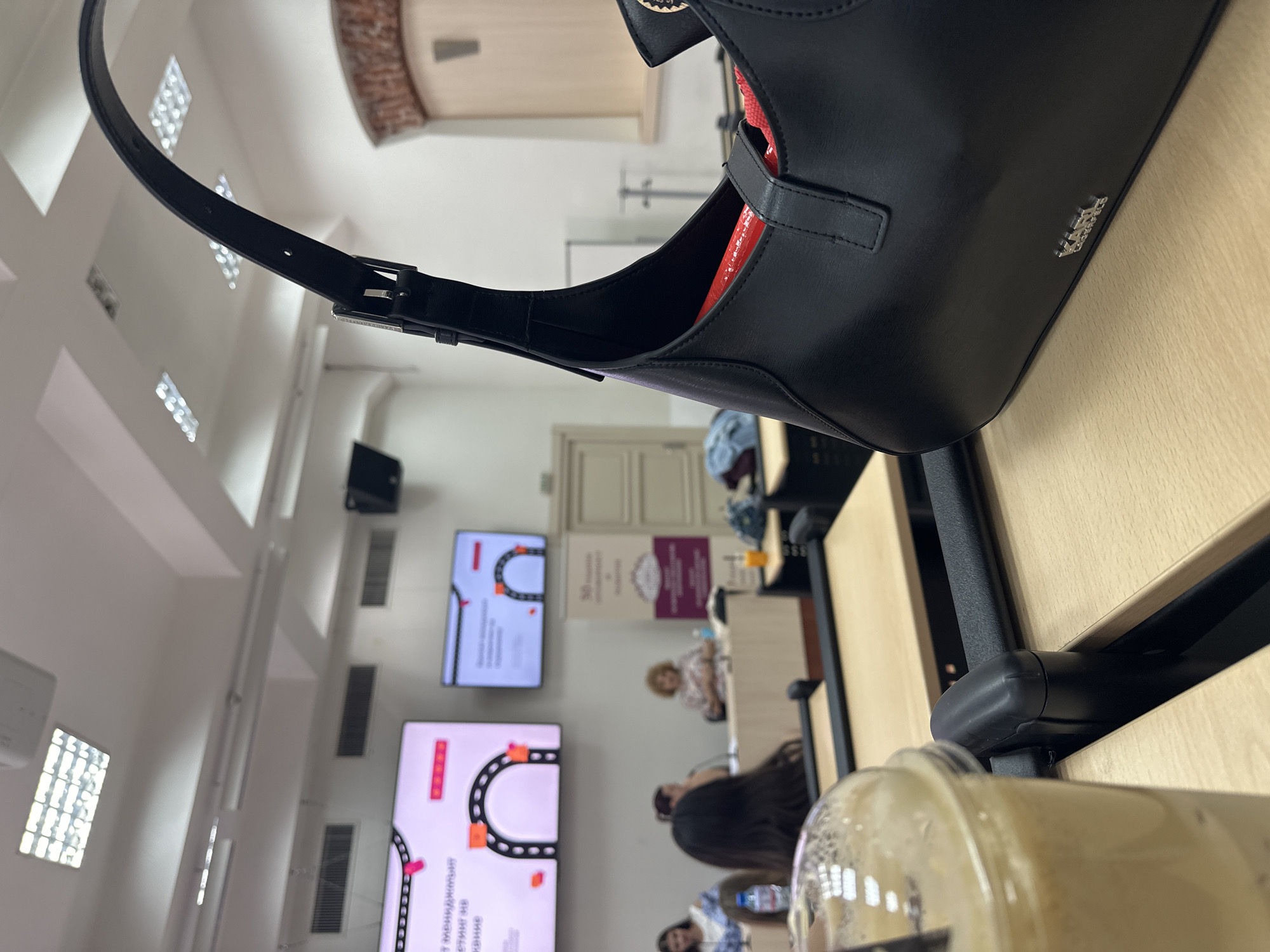 The image depicts an indoor scene, possibly a lecture hall or meeting room, in Sofia, Bulgaria. The foreground is dominated by a black handbag, obscuring part of the scene. In the background, two display screens feature presentations, and several people are seated at tables. The environment seems to be an academic or professional setting.