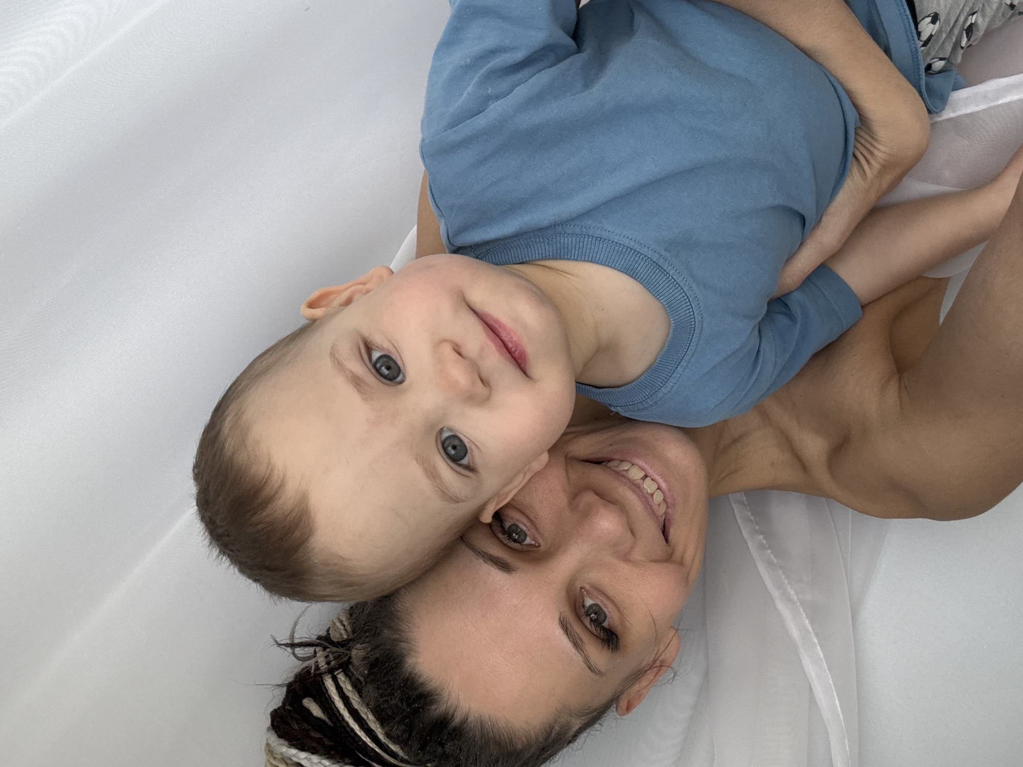 A mother and child are lying on a white surface, likely a bed or blanket. The child, wearing a blue shirt and patterned pants, is nestled against the mother, who is smiling. The mother's hair is styled in braids. The background appears to be a white curtain or draped fabric. There is no information in the image to identify the location as Rusanovo, Russia.