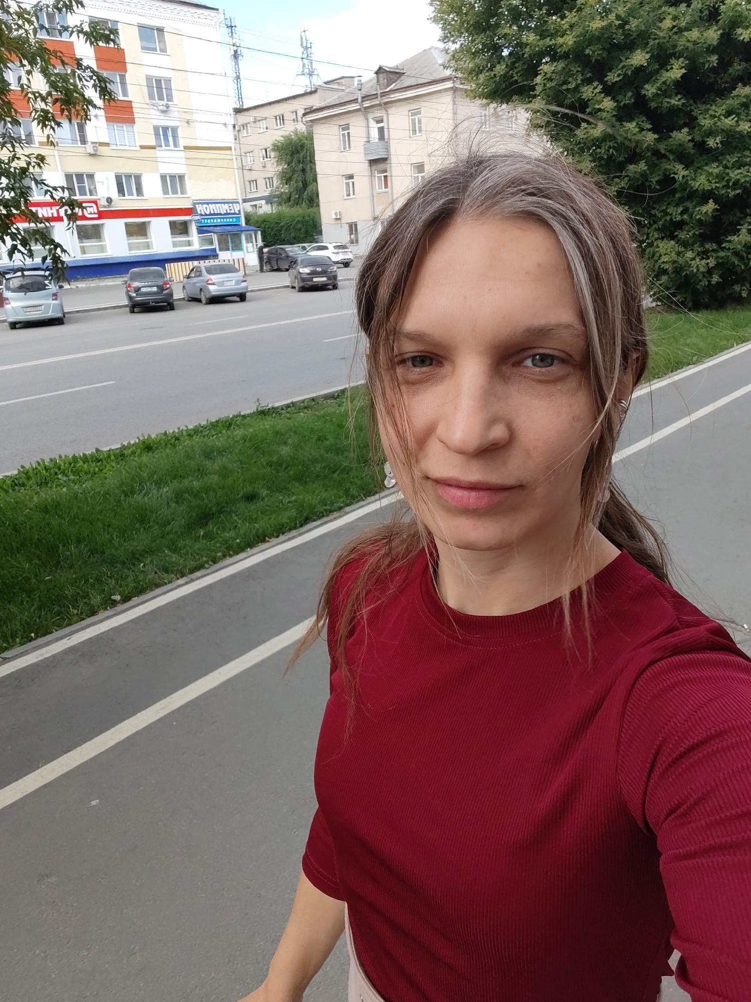 A woman with blonde and graying hair, pulled back into a ponytail, stands on a paved path next to a grassy median. She is wearing a dark red ribbed t-shirt. She is looking towards the camera and smiling slightly. Behind her, a street with several parked cars is visible, as well as a few buildings. The buildings appear to be in an urban setting. The sky is overcast.
