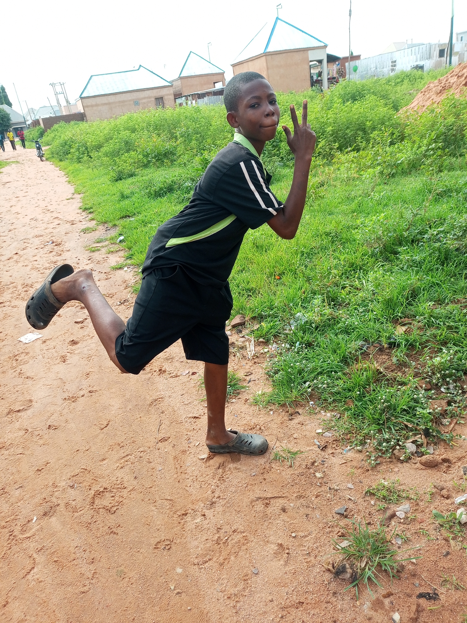 A young boy, appearing to be of Nigerian descent and of early adolescent age, is captured in a full-body, low-angle shot, standing on a sandy path. He is wearing a black t-shirt with a lime green trim and black shorts. His left leg is bent at the knee, raised behind him, while his right leg supports his stance. He is wearing dark-colored slip-on shoes. The boy is looking towards the camera, with his mouth slightly open and his right hand held up in a peace sign gesture, with his index and middle fingers extended.
The environment suggests a rural or suburban setting, likely within the city of Tempure, Nigeria. A dirt path, characterized by its sandy texture and scattered debris, occupies the foreground and extends into the mid-ground. To the right of the path, a dense growth of green grass and low-lying vegetation thrives. In the background, several structures are visible. These include buildings with light-colored walls and distinctly angled, light blue roofs. A motorcycle is parked on the path in the mid-ground, further suggesting a human presence in the area. The sky above is overcast. The overall impression is a candid moment captured outdoors in a simple, possibly developing, community.