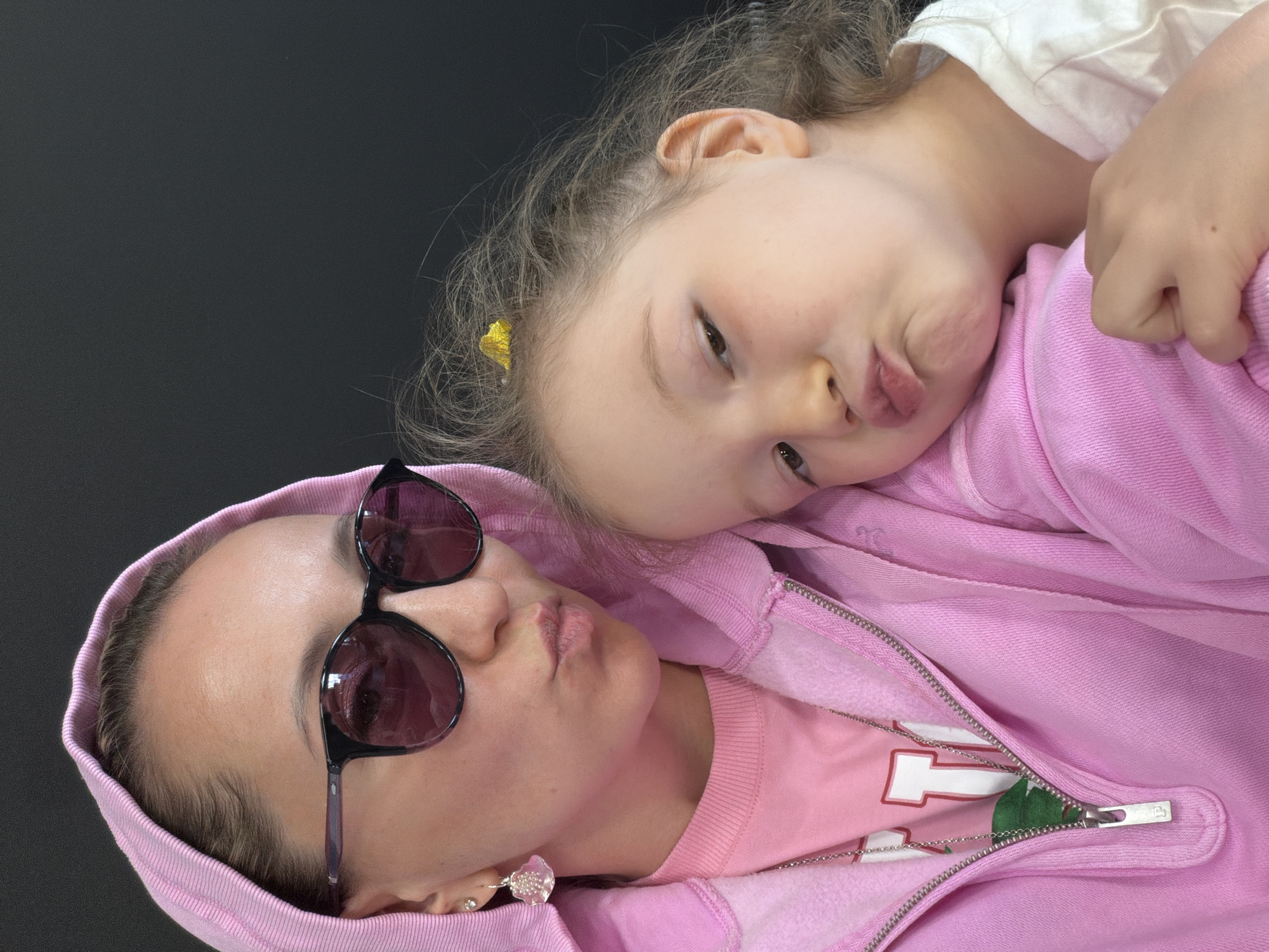 A fair-skinned woman and child are lying down next to each other. The woman wears sunglasses and a pink hooded sweatshirt, zipped open enough to show a pink t-shirt underneath, plus a necklace and earrings. A fair-skinned child with light brown hair is lying with her head resting on the woman's sweatshirt. The background is a plain dark gray. The location is unknown.