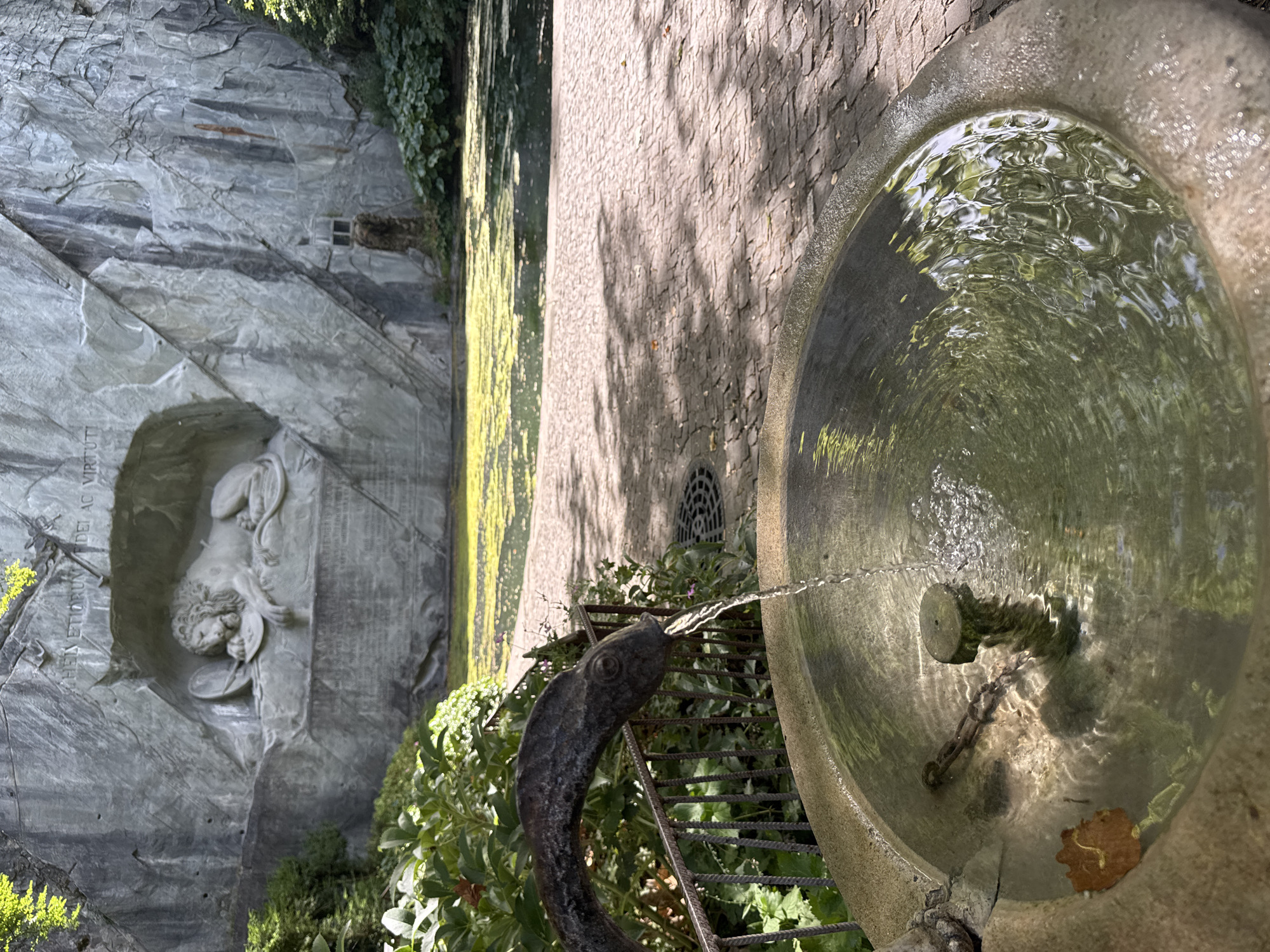 The image depicts the Lion Monument in Lucerne, Switzerland, a famous rock relief carved into a sandstone cliff face. In the foreground, a stone fountain is visible, with water flowing from a sculpted fish-shaped spout into the basin. The water in the fountain is clear and rippling. Behind the fountain, a paved walkway made of brick leads towards a pond covered with green algae. The Lion Monument itself is a prominent feature of the scene, showing a dying lion carved in relief, appearing to be protecting a shield. The monument is set within a rock face, with some text inscribed above it. Lush green vegetation surrounds the monument and the pond. The overall environment appears to be a park or garden area, with dappled sunlight filtering through the leaves of trees, casting shadows on the walkway. No people are visible in the image.