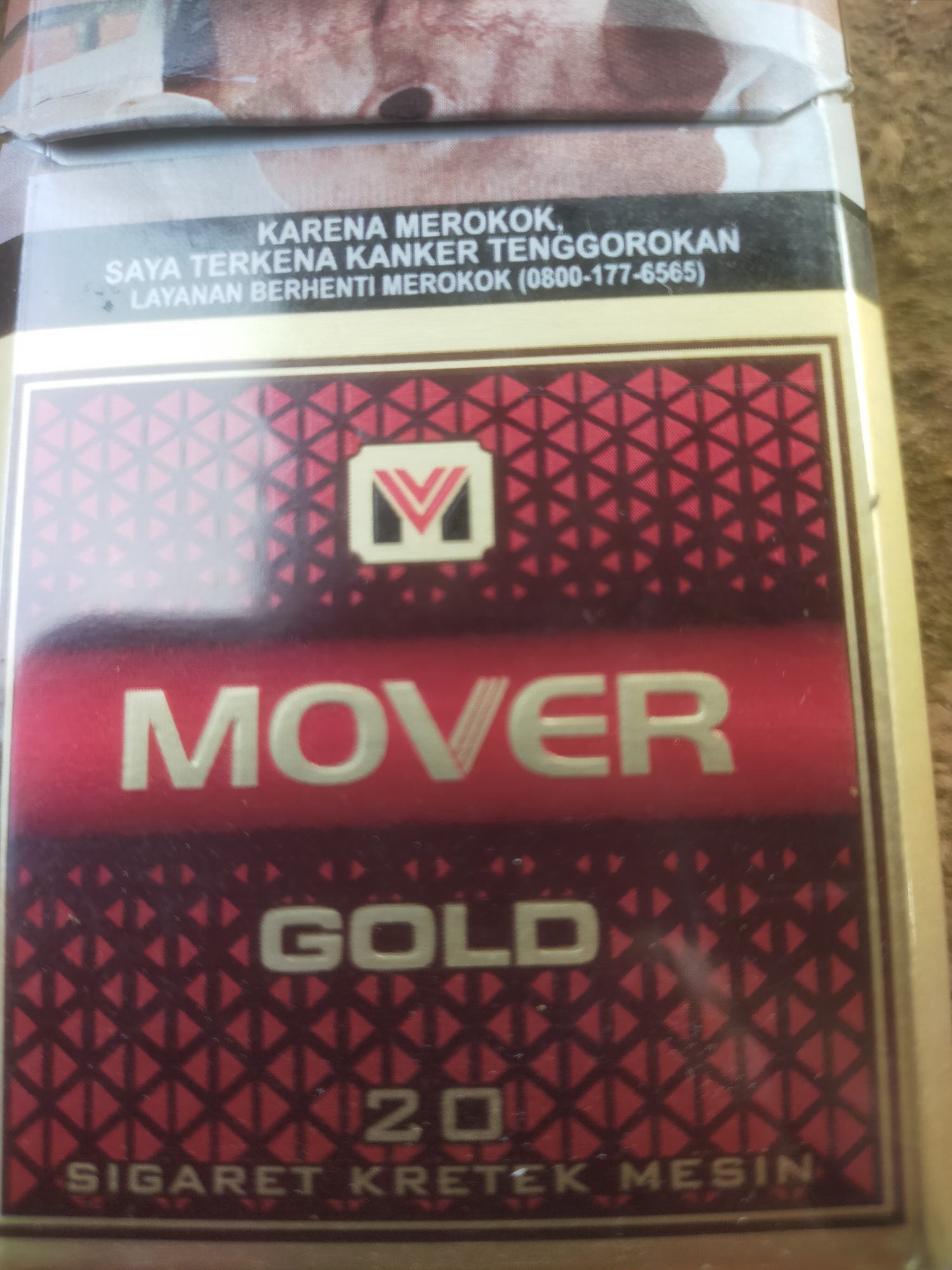 The image is a close-up of a cigarette pack. The pack is predominantly red and gold. The top of the pack features a black band with white text, which appears to be a warning message. Below this band, there is a metallic red rectangular area containing the word "MOVER" in large, bold, gold lettering. Beneath "MOVER," the word "GOLD" is also written in gold. Further down, "20" is visible, followed by "SIGARET KRETEK MESIN" in smaller gold lettering. The background of the pack is a red geometric pattern. The image is framed with a golden border. The lighting suggests an indoor setting, possibly a retail environment, though no specific location within Padang Dalom, Indonesia, can be determined from the visual information alone.