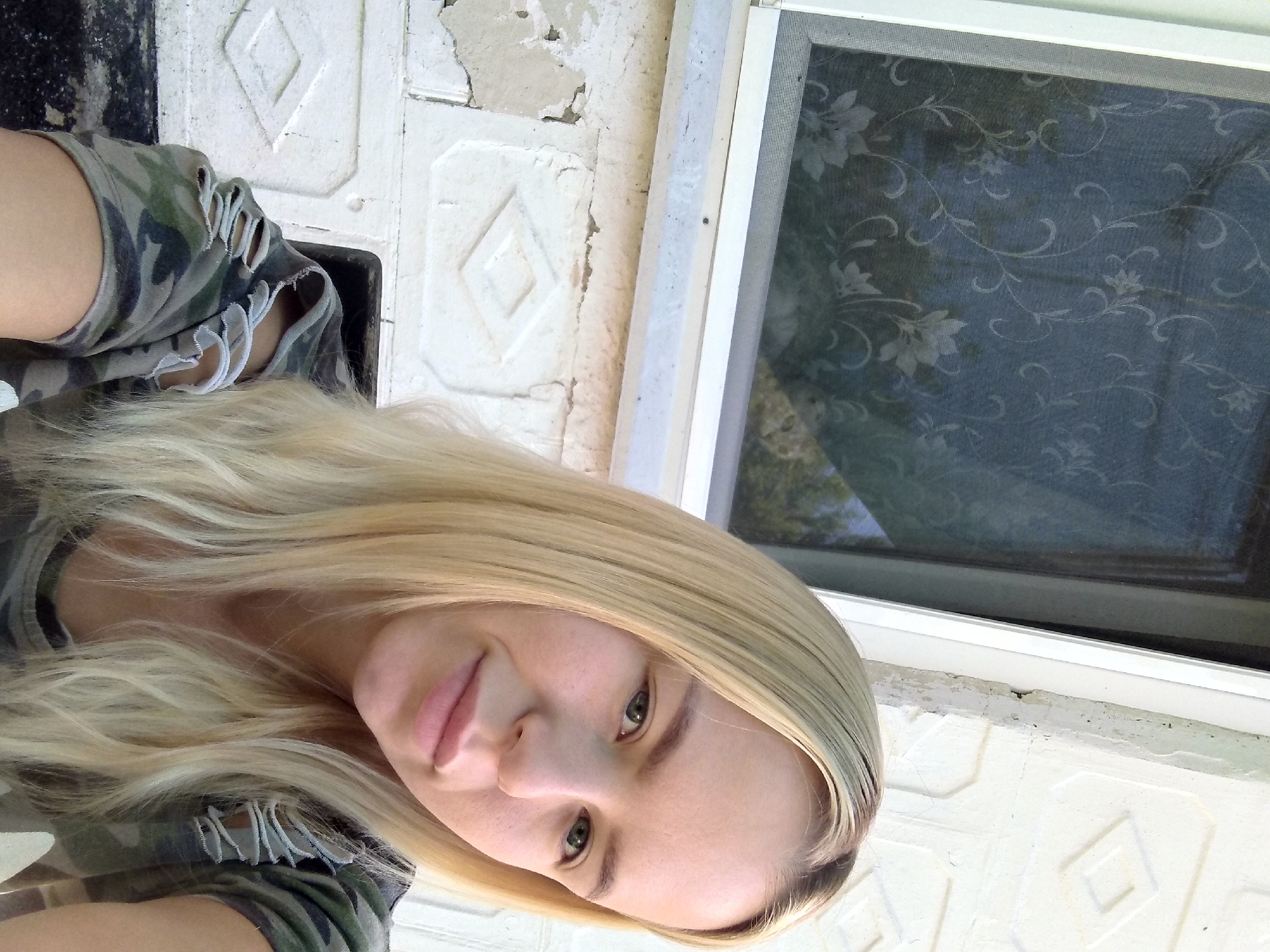 In Radisne, Ukraine, a young woman with blonde hair takes a selfie outdoors. She is positioned in front of a white brick wall with a diamond pattern and a window with a floral screen. The woman is wearing a camouflage shirt with ripped sleeves. The lighting suggests it is daytime, with sunlight casting shadows.