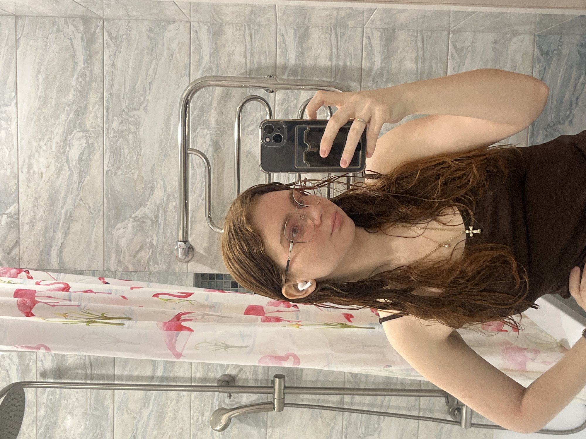 The image shows a young woman taking a mirror selfie in a bathroom. She has long, wavy, wet brown hair, suggesting she may have recently showered. She wears round, thin-framed glasses and a white AirPod in her left ear. Her expression is neutral, with a slight pout. She is dressed in a dark brown tank top and accessorized with two necklaces: one featuring a small gold cross pendant and another with a pearl. On her right hand, which holds a dark smartphone with a clear case, a silver ring is visible on her ring finger.
The bathroom, located in Kazan, Russia, features walls covered in light-colored, grey and white marbled rectangular tiles. A silver, wall-mounted heated towel rail is positioned behind her head. Below her, a white and pink shower curtain adorned with flamingo patterns is partially visible, indicating a shower area. A shower head and hose fixture are seen at the bottom left of the frame, and the edge of a white bathtub or shower base is visible on the right. The indoor lighting suggests it is likely daytime. No visible text is present in the image.