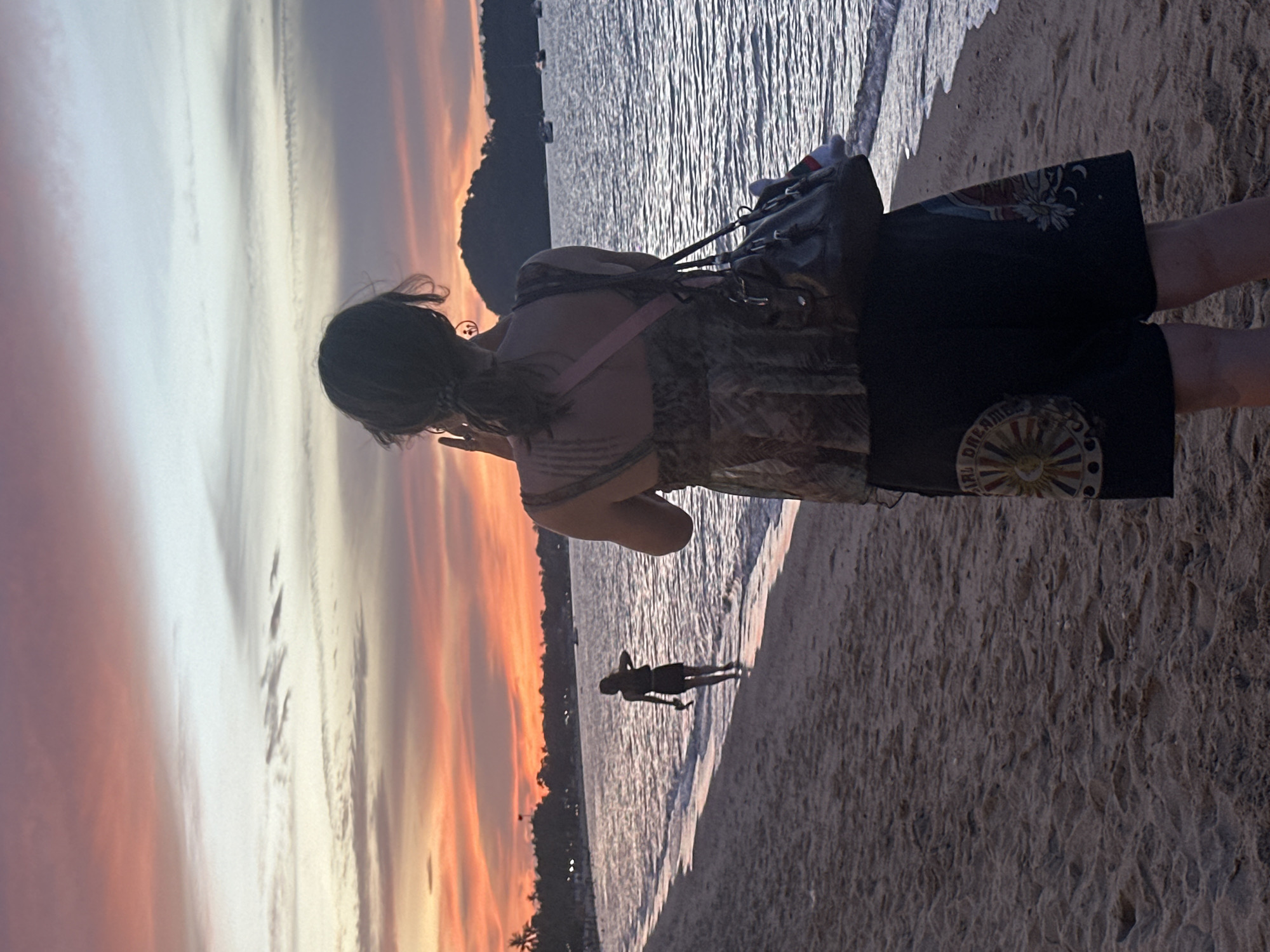 A person with dark hair pulled back stands on a sandy beach, facing the ocean and a colorful sunset. They are wearing a patterned top and dark shorts with embroidered designs. The person is holding a phone up, appearing to take a picture of the sky. A dark bag with straps hangs from their shoulder. Further down the beach, another person in a dark outfit is walking near the water's edge, their back to the viewer. The ocean is calm with gentle waves lapping the shore, and several boats are visible in the distance. The sky is filled with hues of orange, pink, and purple, indicating sunset. Palm trees and a silhouetted landmass can be seen along the coastline. The scene suggests a serene evening on a beach in the Ko Samui District of Thailand.