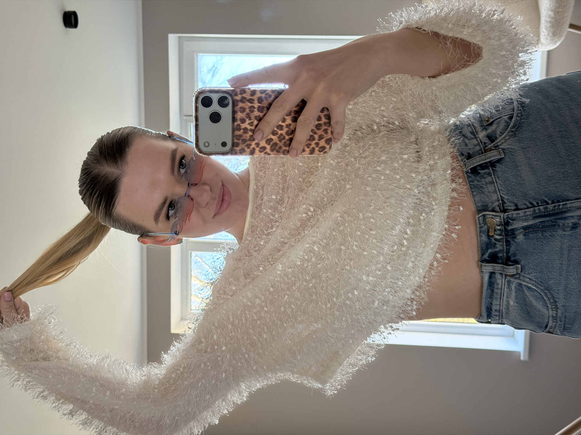A young woman is depicted taking a mirror selfie, with the image rotated 90 degrees clockwise. She is positioned diagonally across the frame, looking towards her reflection in the mirror with a subtle smile. Her light-colored hair is styled in a high ponytail, which she holds with her left hand. She wears distinctive flame-shaped sunglasses with pinkish, semi-transparent lenses.
Her outfit consists of a cropped, long-sleeved top made from a fuzzy, off-white or cream-colored material that reveals her midriff. She pairs this with light blue denim jeans. In her right hand, she holds a smartphone, clad in a leopard print case, aiming it towards the mirror to capture the image.
The background shows an indoor setting with light-colored walls. A large window is visible, through which natural daylight streams in, illuminating the scene. Bare tree branches can be seen outside the window. A dark, circular object is mounted on the wall in the upper left section of the image. The overall impression is bright and casual. There is no visible text in the image. The location context is Dreiliņi, Latvia.