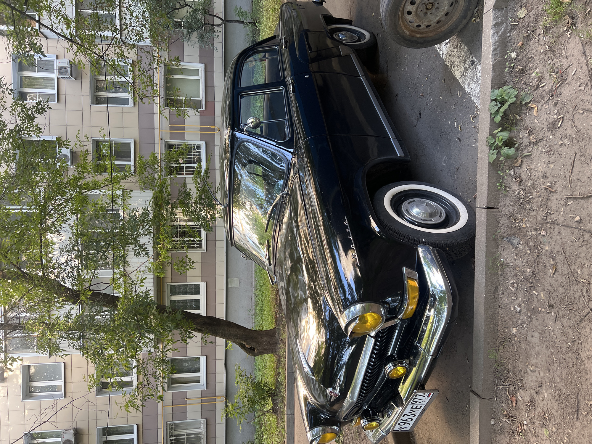 A black vintage sedan is parked on a dirt and asphalt lot in front of a multi-story apartment building in Moscow, Russia. The car is a GAZ-21 Volga, identifiable by its distinctive grille, round headlights, and chrome accents. It has white-wall tires and a license plate reading "K963ME 977 RUS". The apartment building has a beige and brown tiled facade with numerous windows and balconies. Trees with green foliage are scattered around the scene. The ground is a mixture of dirt, gravel, and some patches of grass. A spare tire is visible on the right side of the frame. The lighting suggests it is daytime, with sunlight reflecting off the car's polished surface.