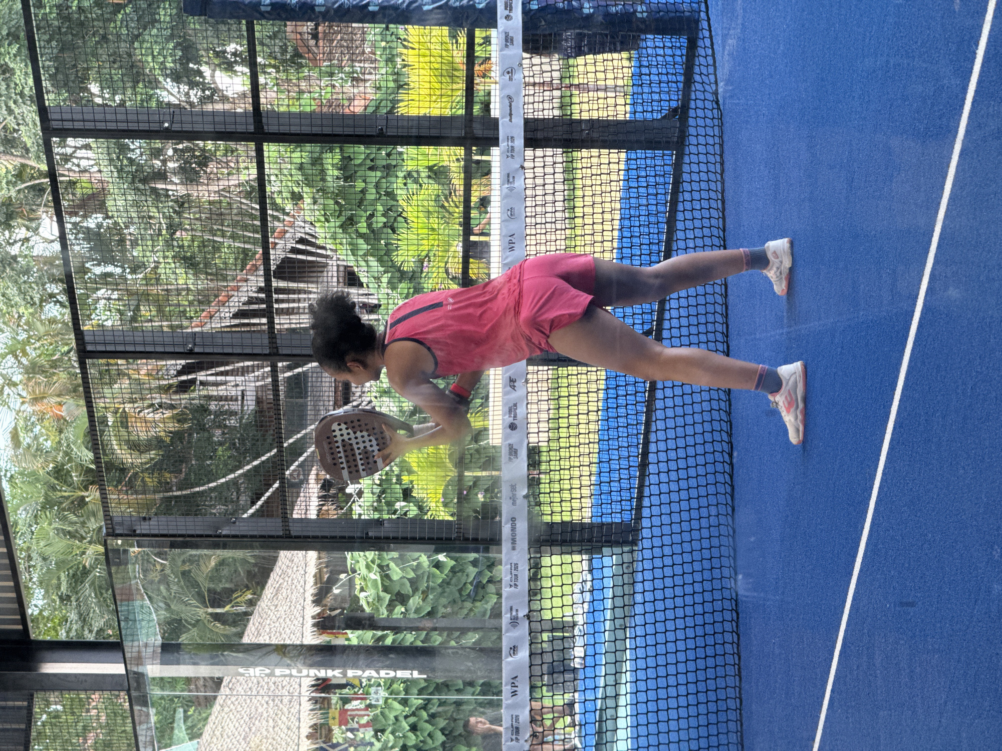 In บ้านบางรักษ์, Thailand, a young person with dark, curly hair, wearing a pink sleeveless athletic top and pink shorts, stands on a blue padel court. They are holding a brown padel racket, poised to strike a ball. The court is enclosed by glass walls and black metal fencing, with lush green foliage and palm trees visible in the background. A net divides the court. Another person can be seen in the distance on the same court, partially obscured by the glass. Logos for "WPA," "IPPA," "MONDO," and "Better Sek" are visible on banners along the net.